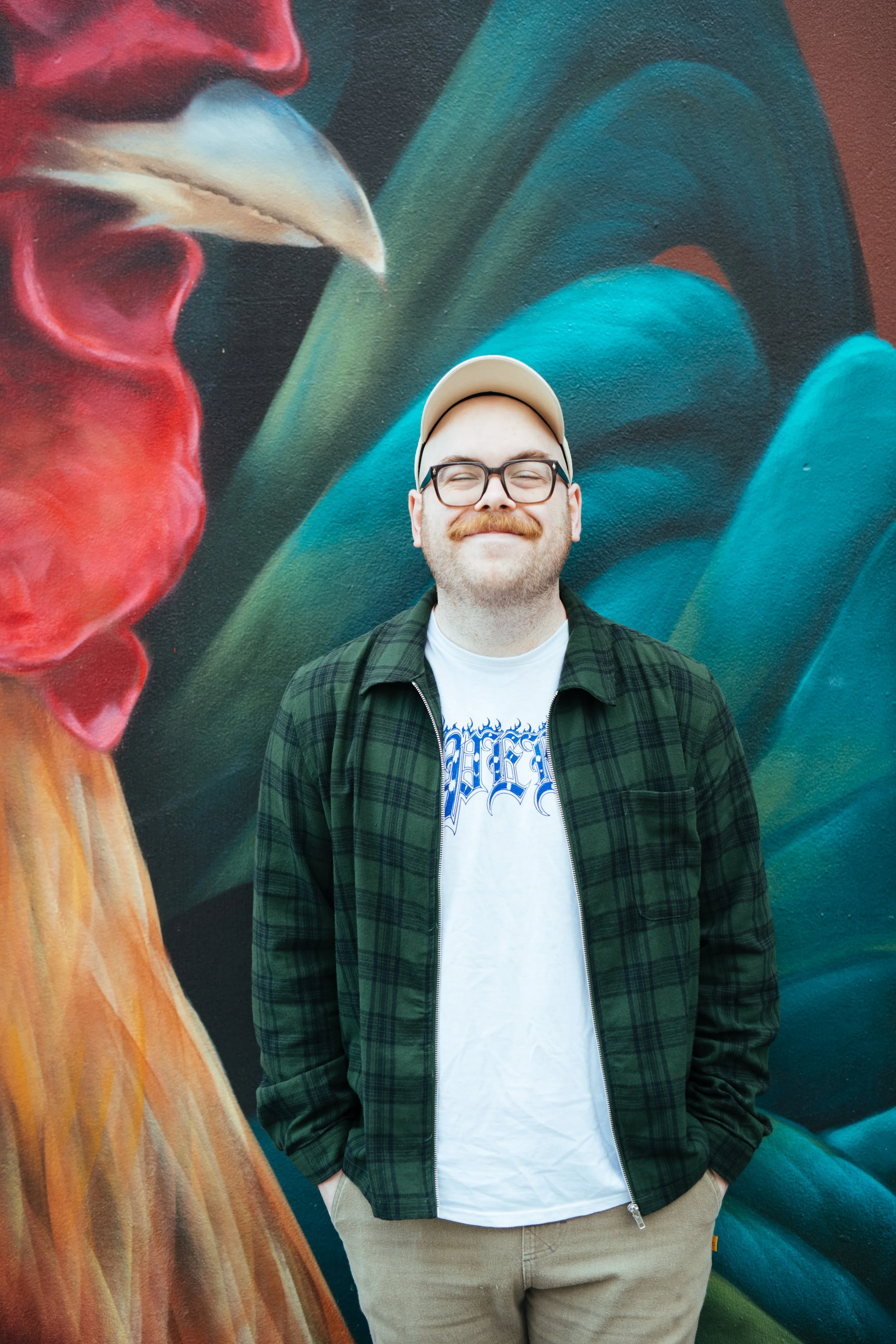 A smiling man with glasses, a beige cap, a green plaid jacket, and beige pants standing in front of a vibrant mural with large leaves and a partially visible face on the left side.