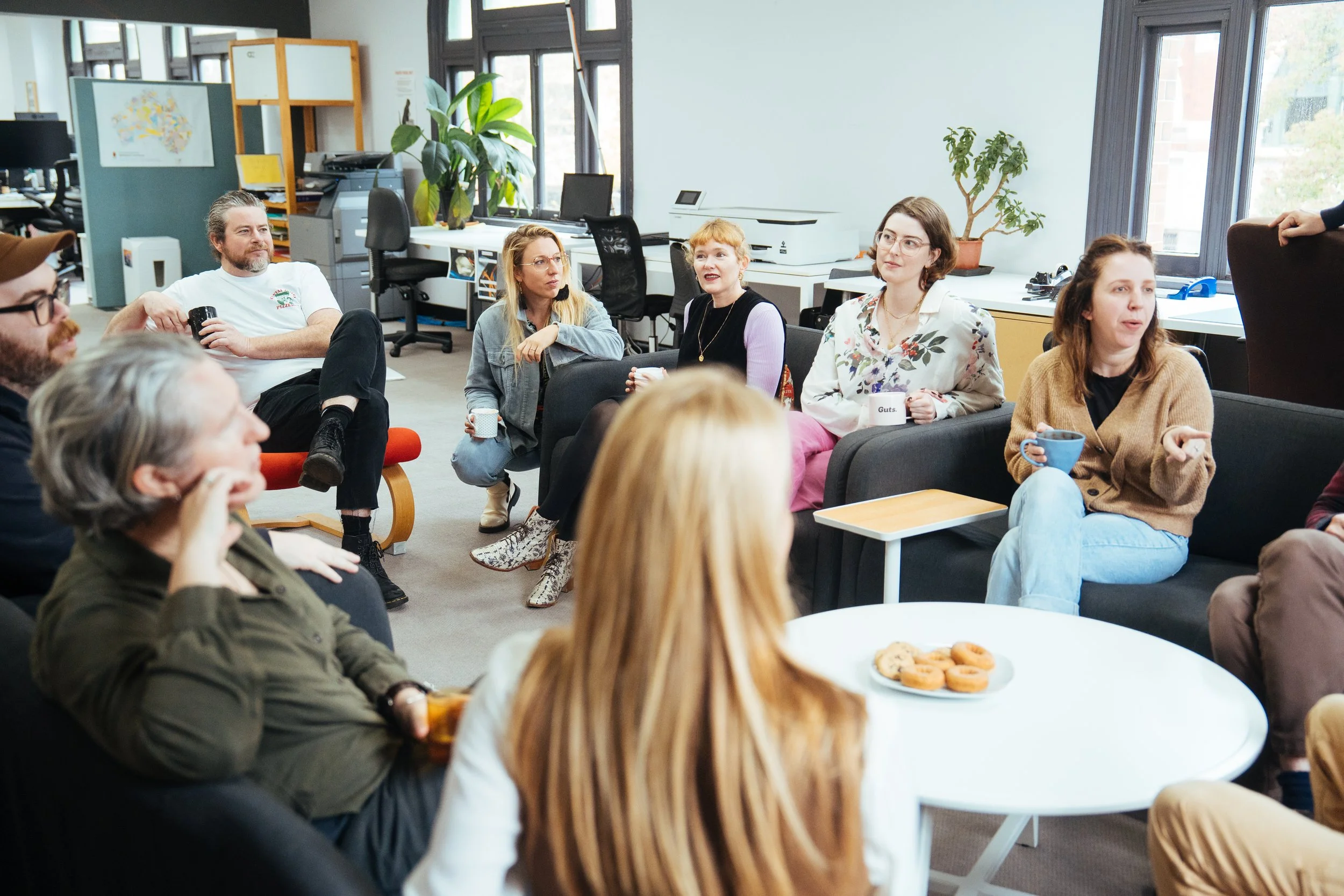A group of people sitting in a modern office, engaged in conversation. Some are holding mugs, with a plate of cookies on a white table in the foreground. Large windows let in natural light, and there are plants and office equipment in the background.