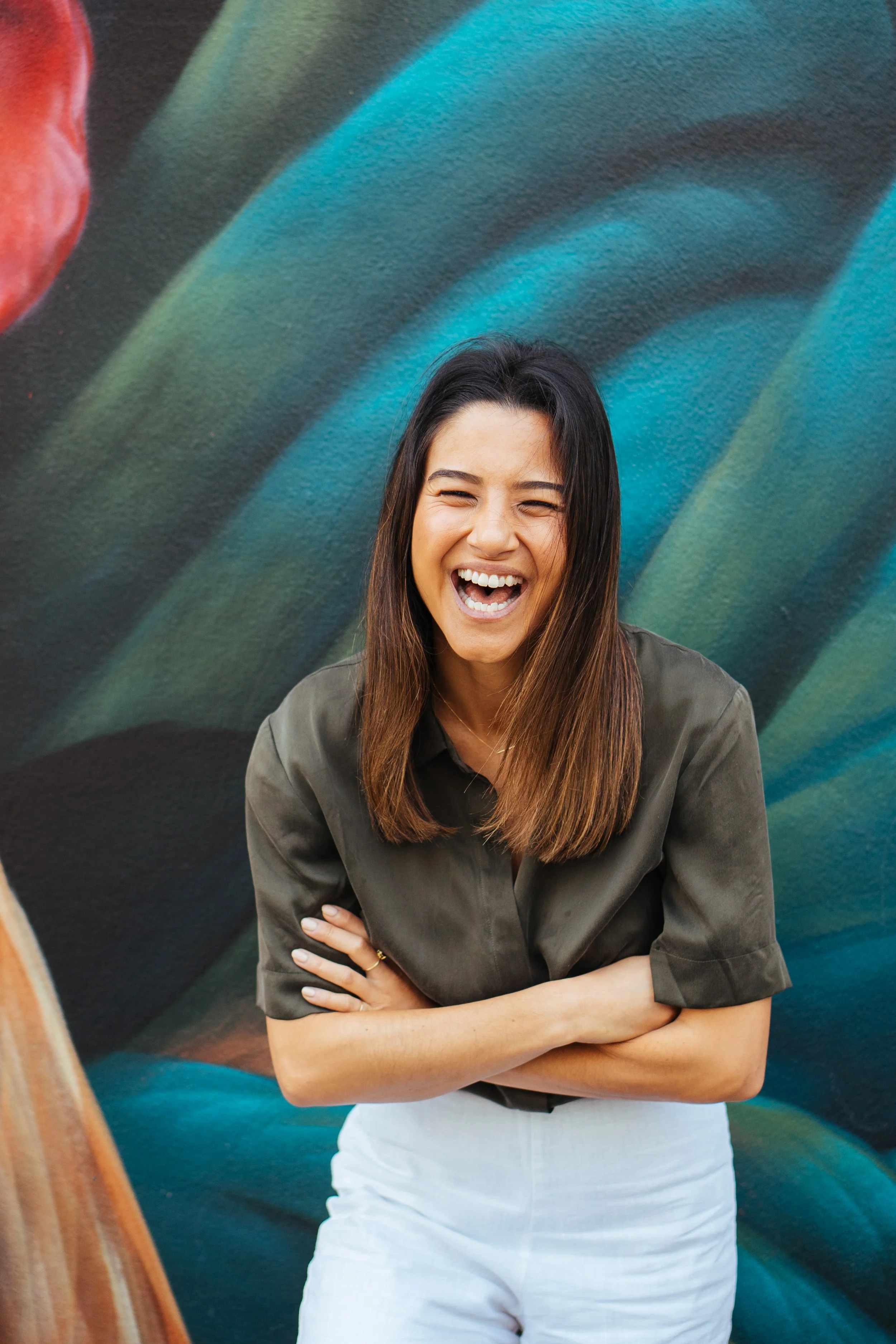 A woman with long dark hair laughing with her arms crossed in front of a colorful mural.