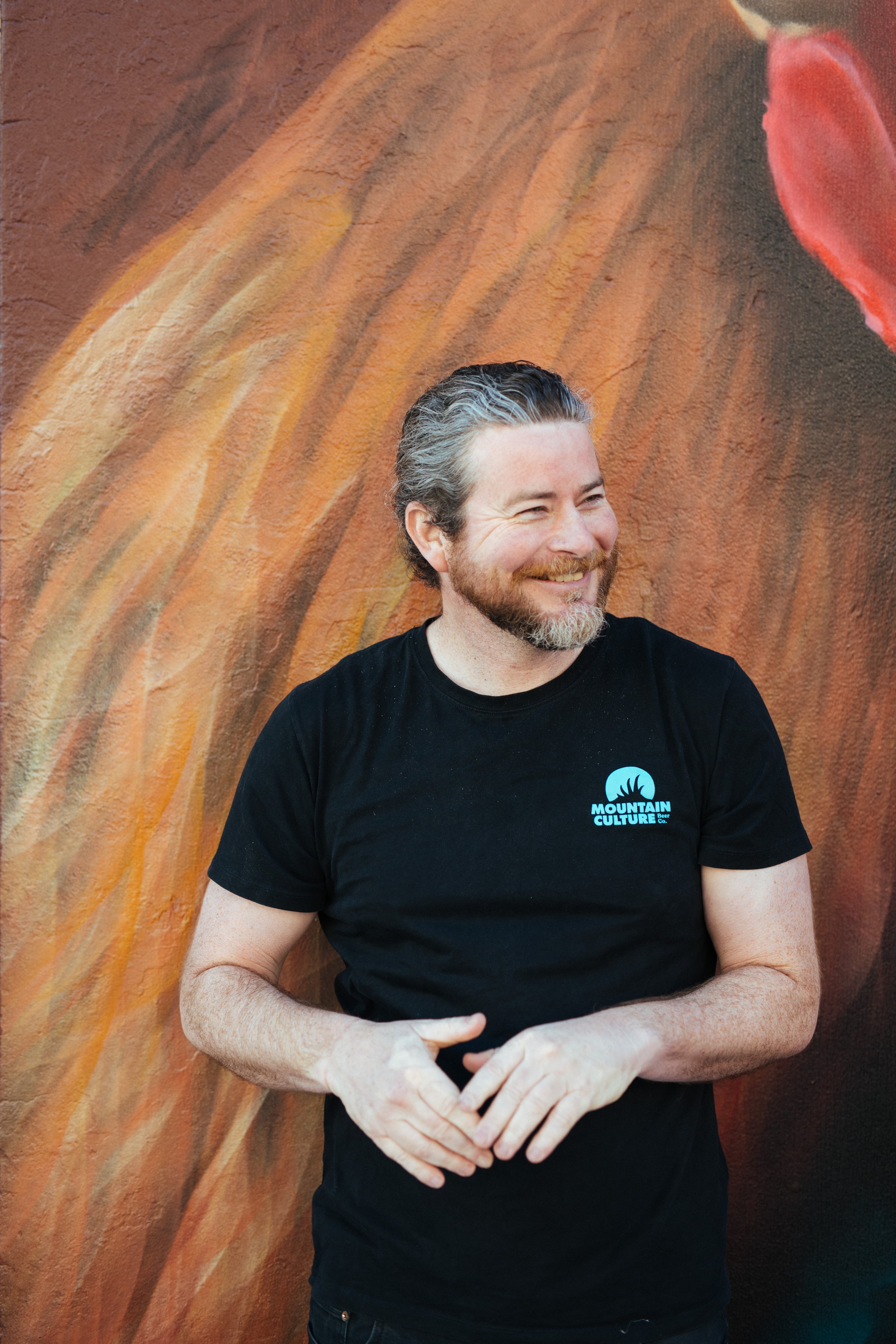 A smiling man with gray hair and beard, wearing a black t-shirt with a "Mountain Culture" logo, standing in front of a colorful abstract mural.