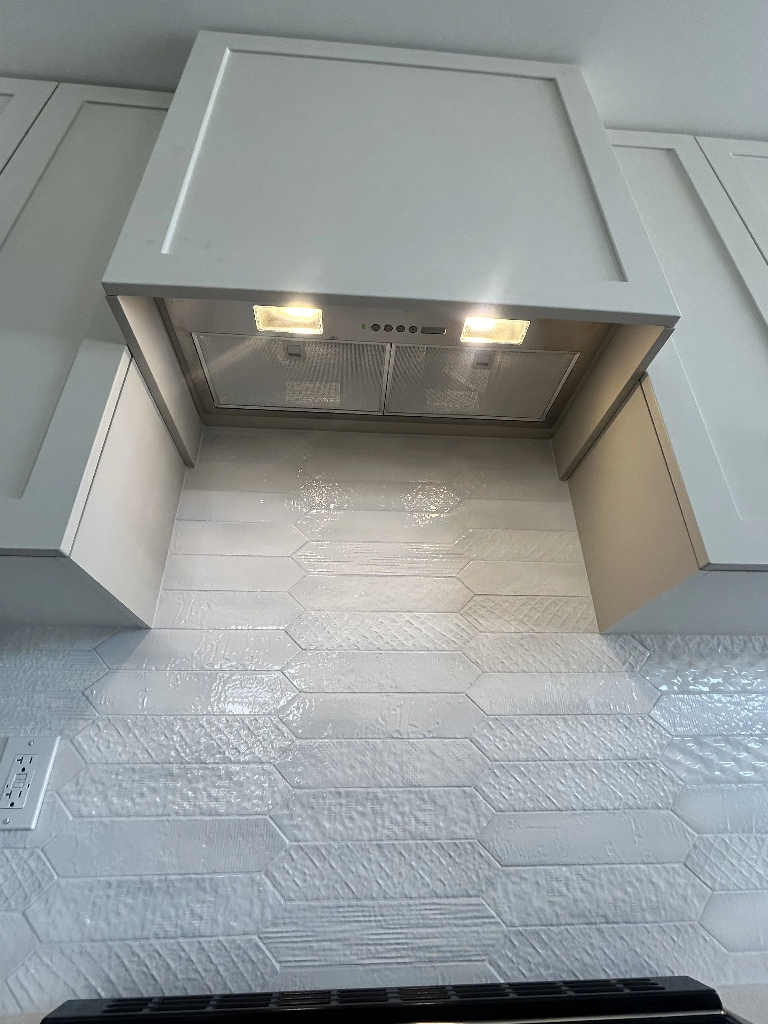 View of a kitchen wall with white tiles and power outlets beneath a vent hood with lights.