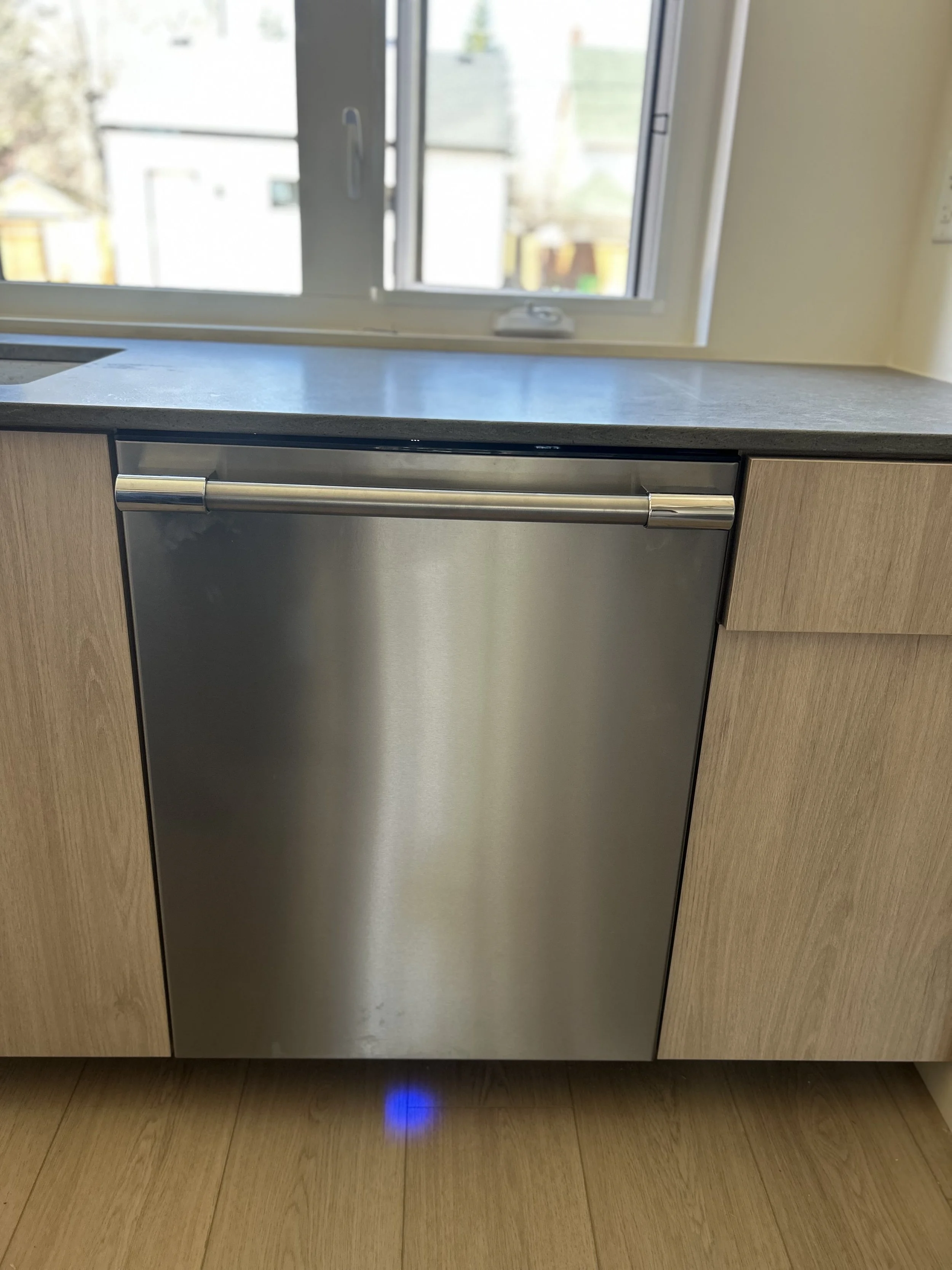 Close-up of a dishwasher installed beneath a light wood kitchen countertop and cabinets, with a view of a window overhead showing an outdoor scene.