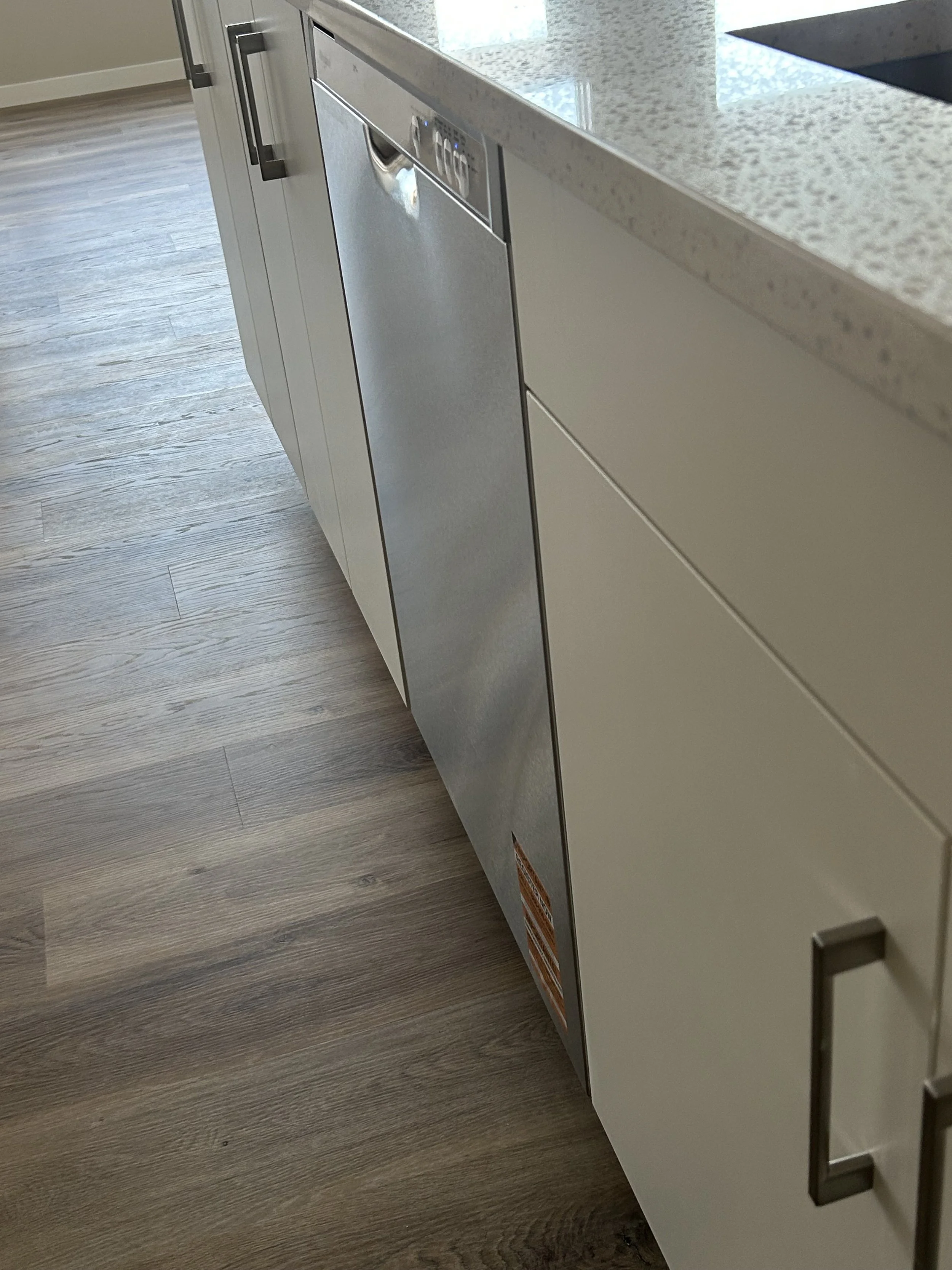 Close-up of a kitchen countertop and dishwasher built into white cabinetry, with light wood flooring underneath.