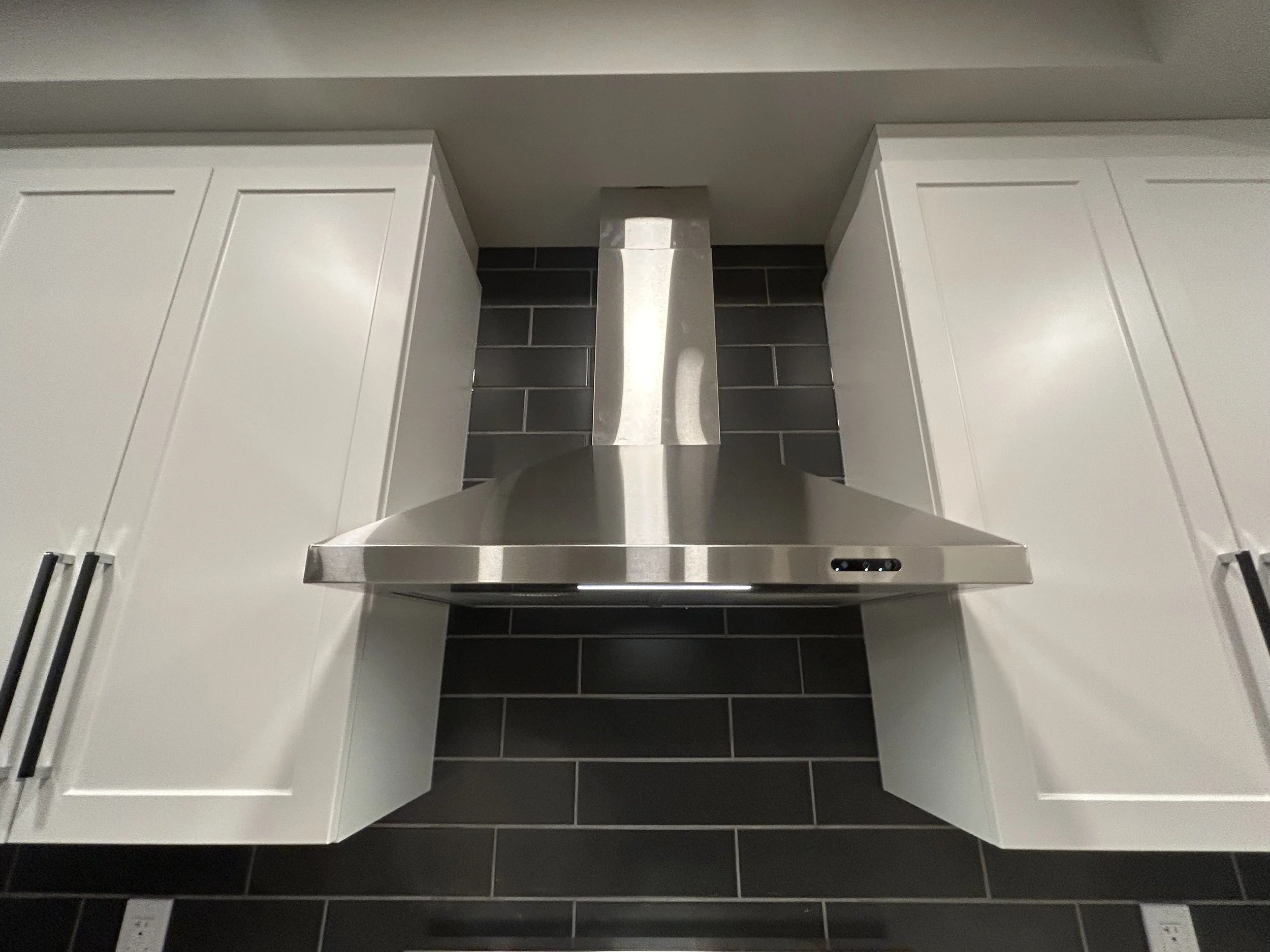 A modern kitchen range hood mounted on a dark tiled wall between white cabinets.