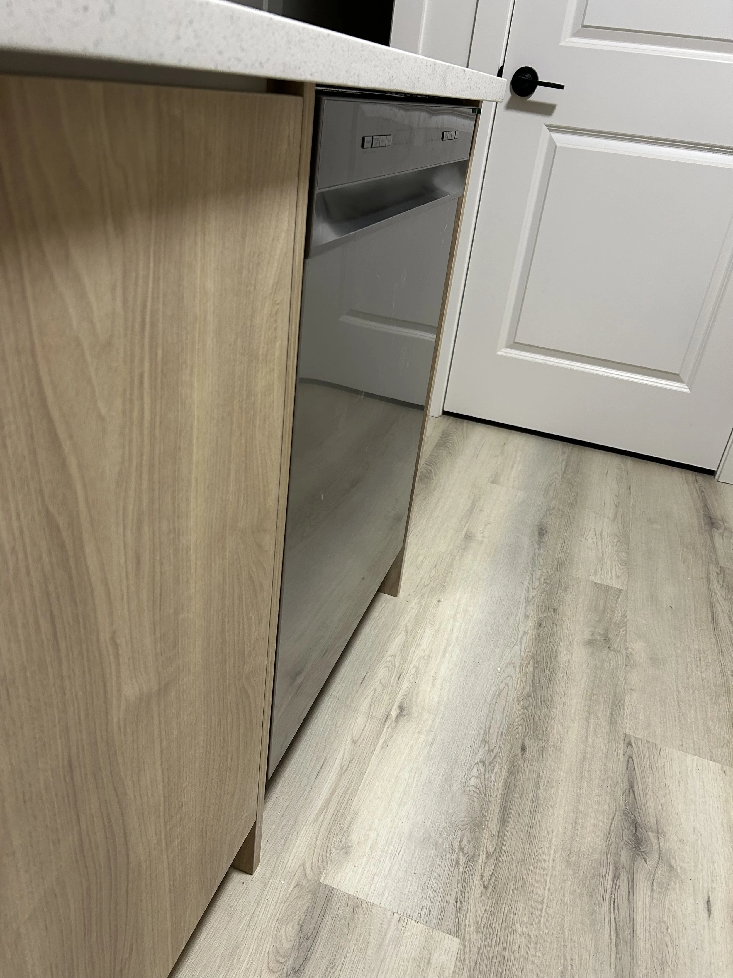 Part of a kitchen with a wooden cabinet, a stainless steel dishwasher, a white countertop, and a white door with black hardware, the floor is light-colored wood.