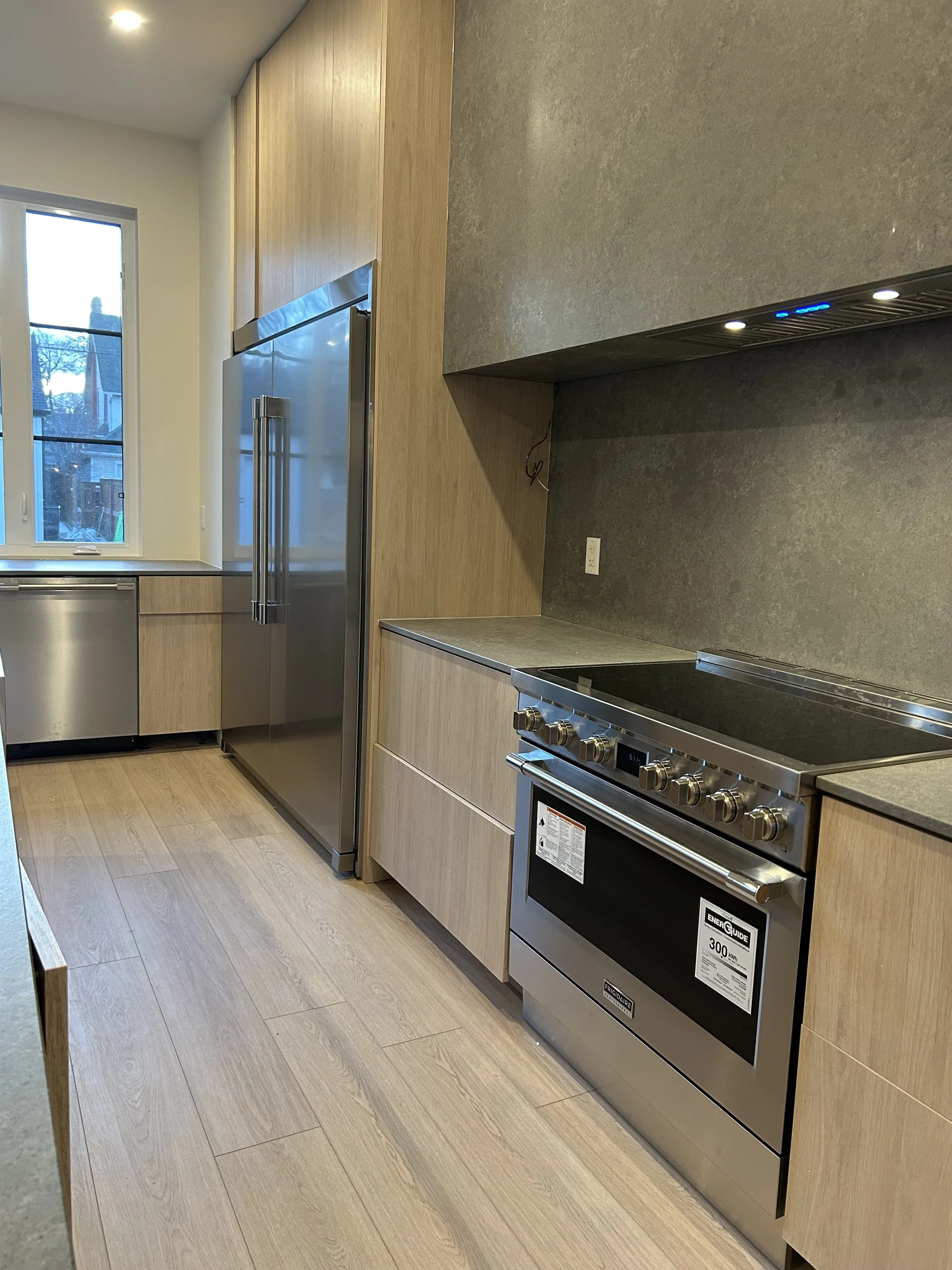 Modern kitchen with light wood cabinets, stainless steel appliances including a large refrigerator and a stove, gray countertops, and wood-look flooring. A window lets in natural light, and recessed lighting is installed in the ceiling.