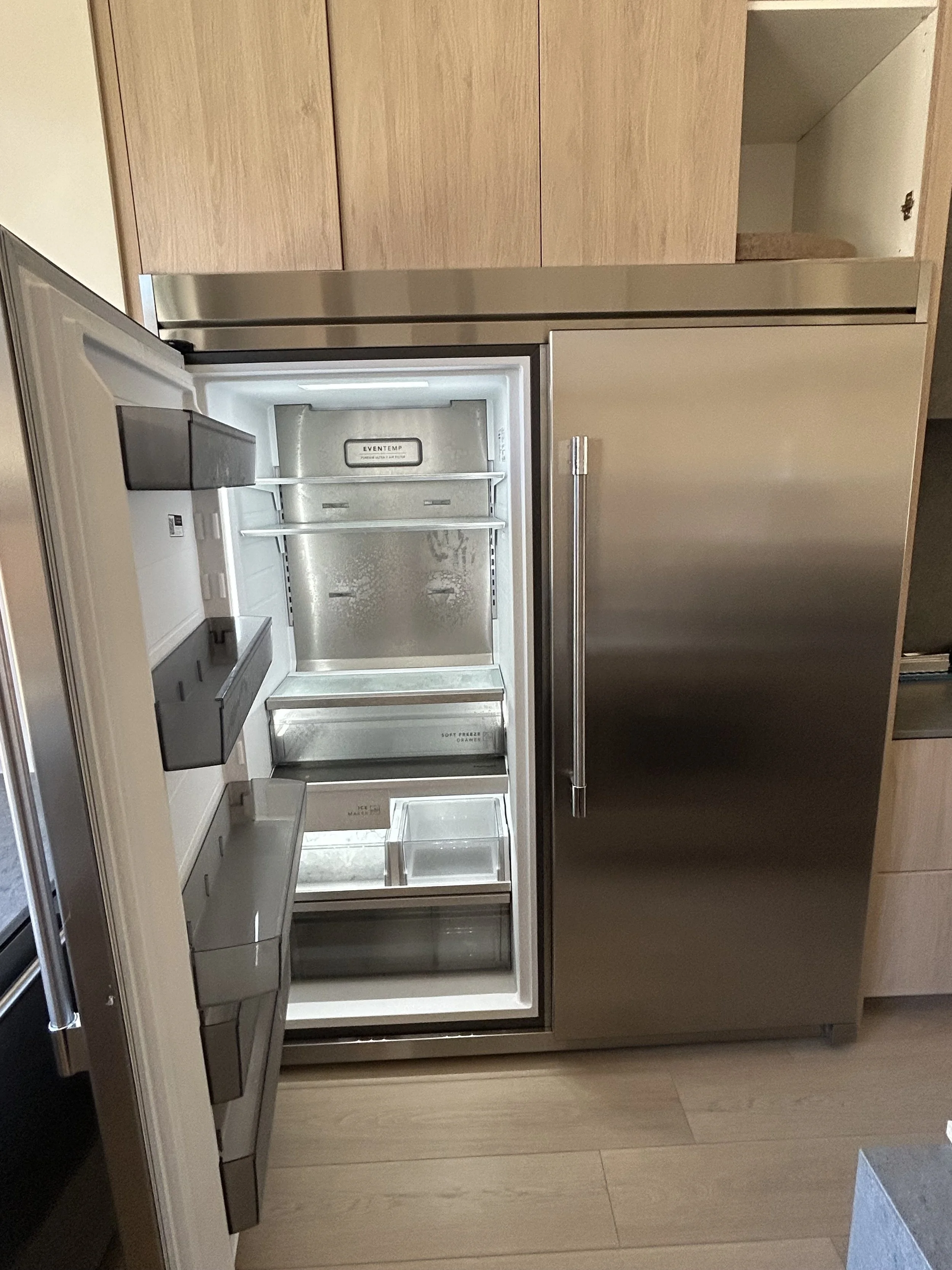 Open stainless steel refrigerator in a modern kitchen with wooden cabinets and a beige floor.
