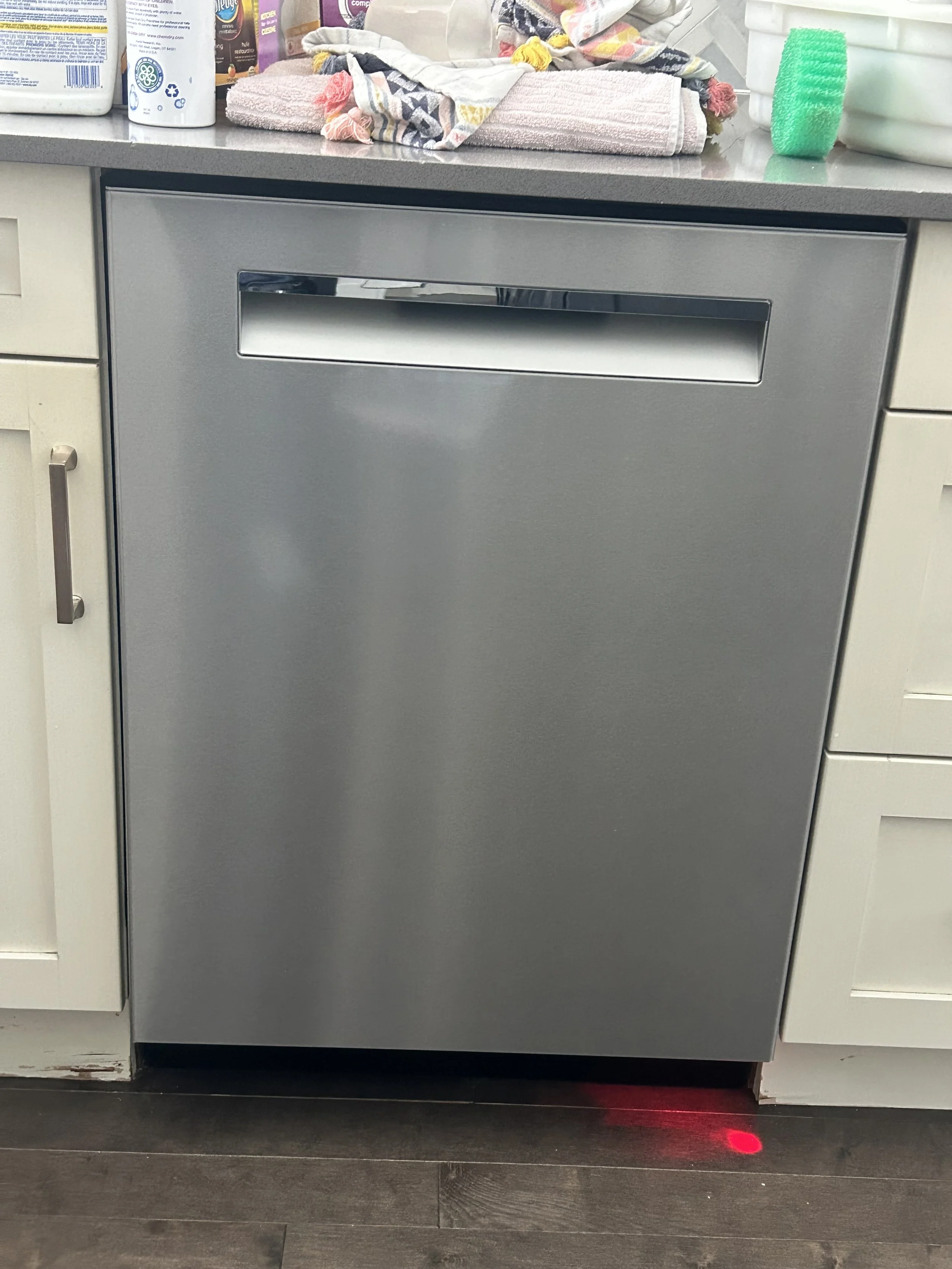 A stainless steel dishwasher in a kitchen, surrounded by various items on the counter including cleaning supplies, towels, and a green scrubber. The floor is dark wood.