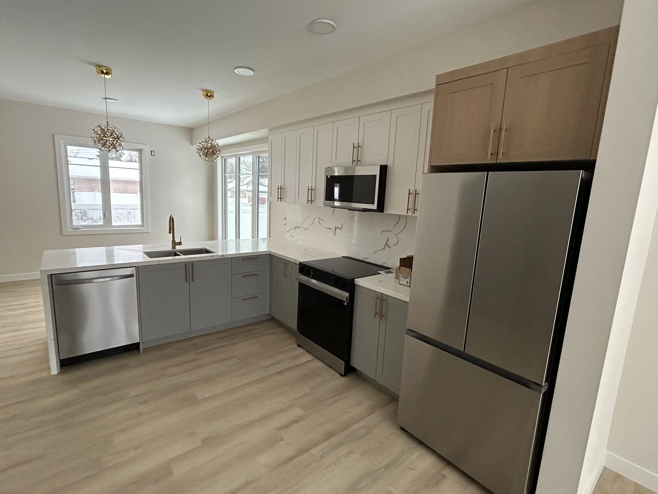Modern kitchen with grey cabinets, marble backsplash, stainless steel refrigerator, and pendant lighting, with windows showing an outdoor scene.