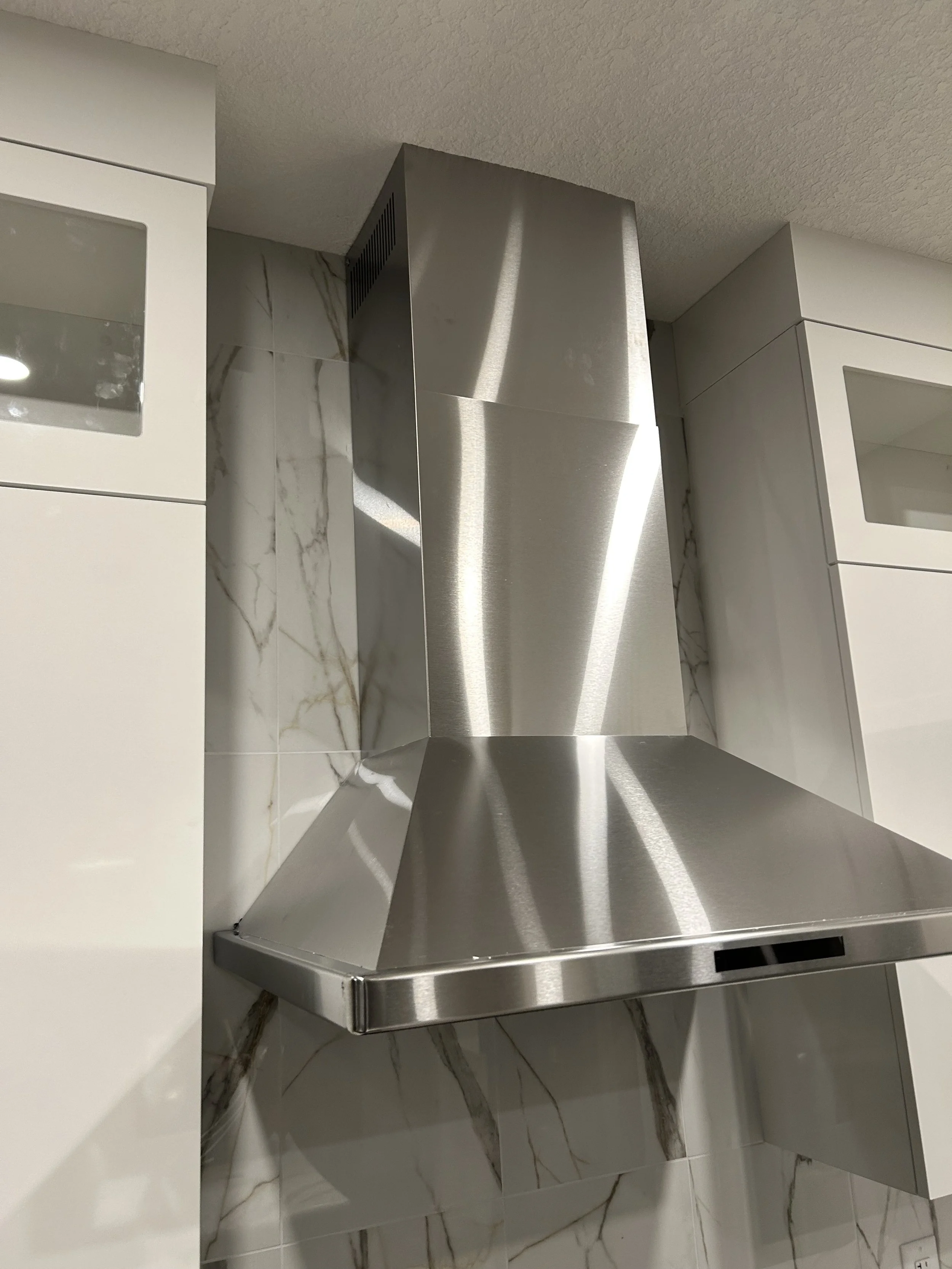 A modern kitchen with a stainless steel range hood above a stovetop, white cabinetry, and marble tile walls.