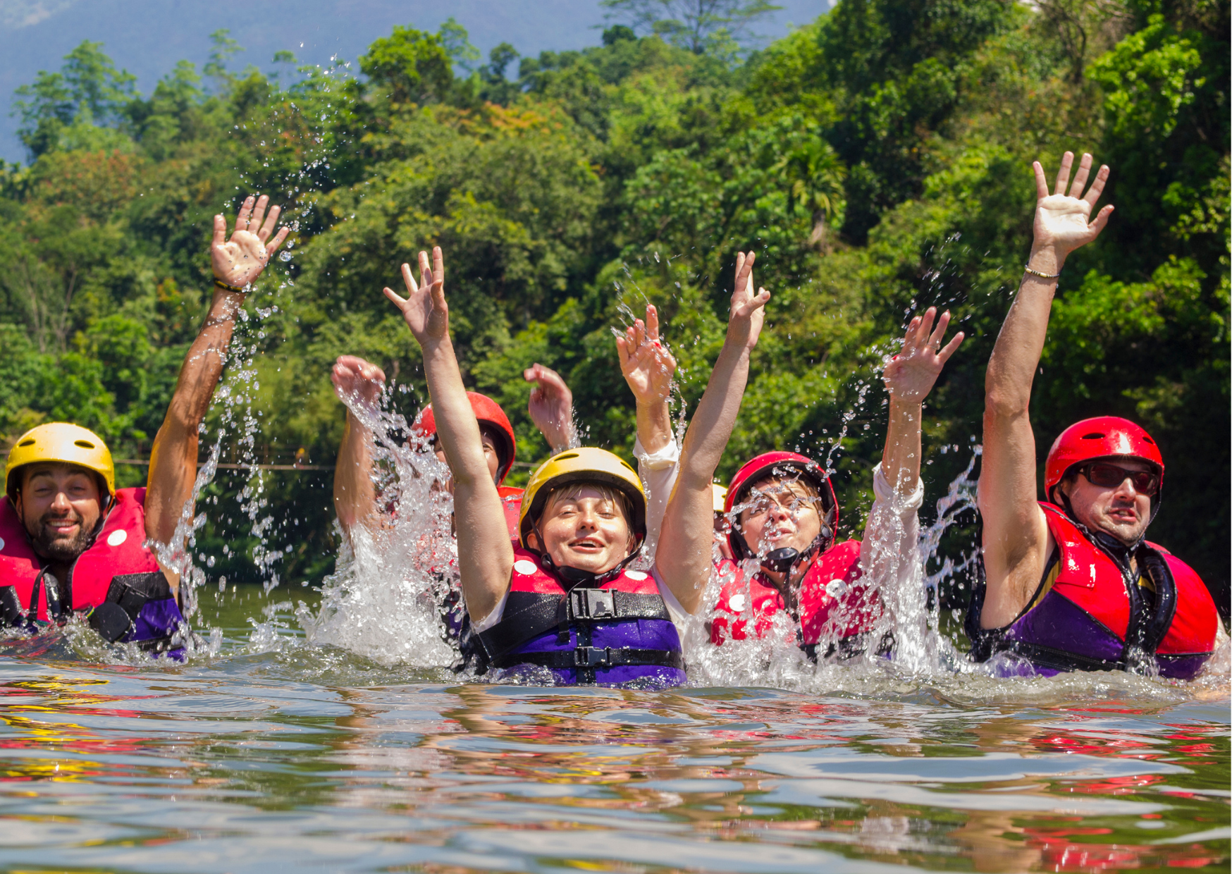 People wearing life jackets and helmets swimming and splashing water in a river surrounded by lush green trees.