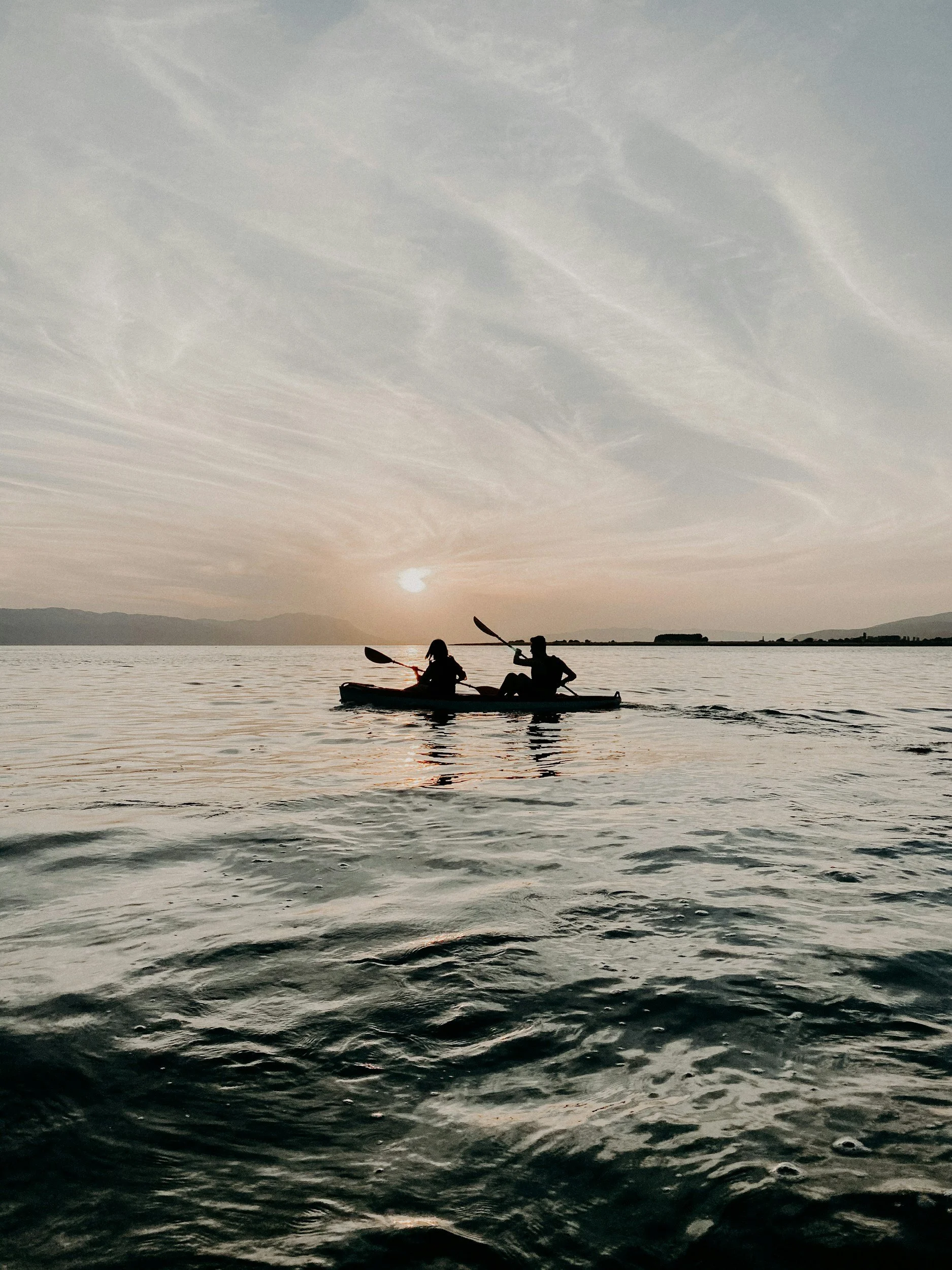 Silhouettes of two people kayaking on a calm lake during sunset with a mountain range in the distance.