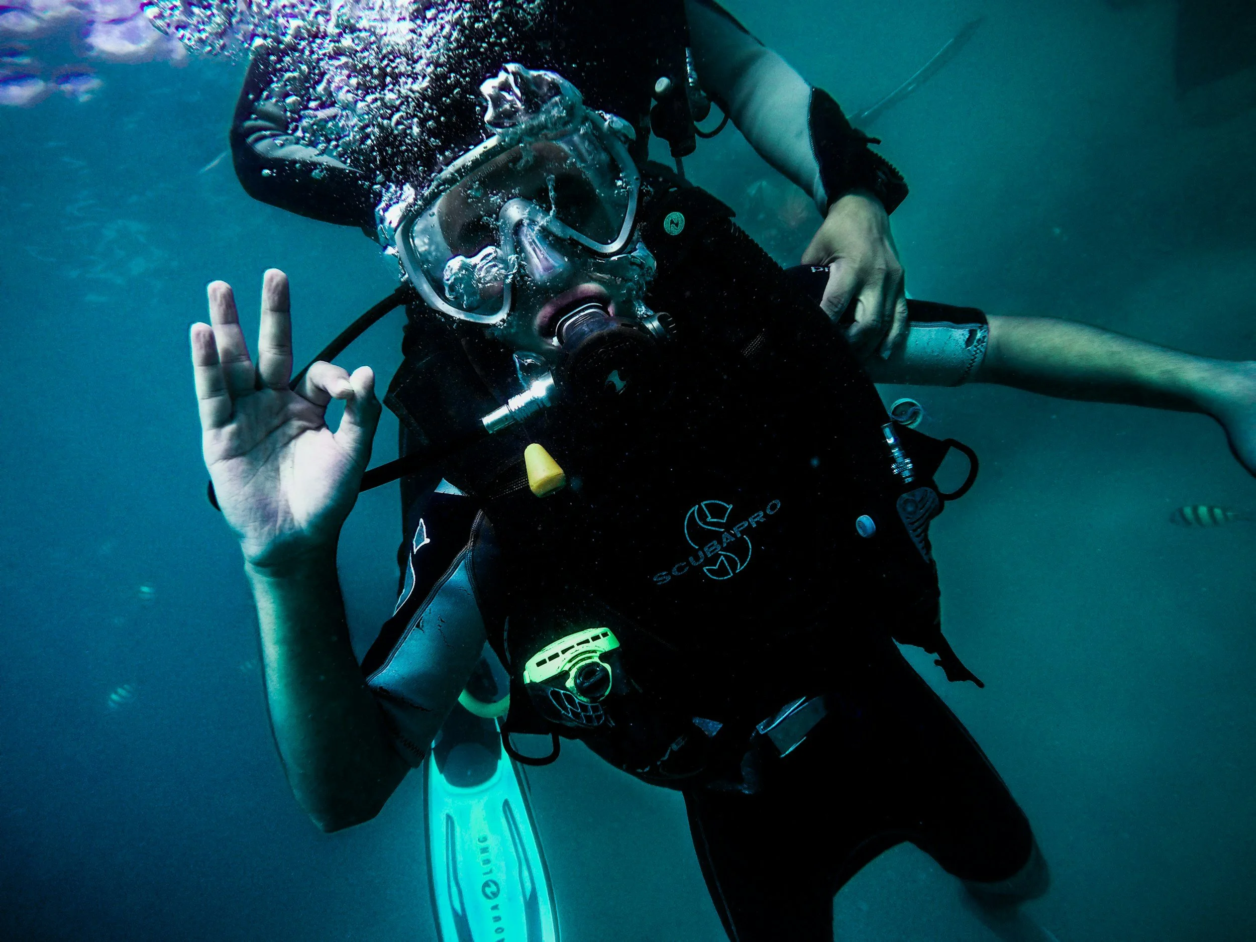 A scuba diver underwater making an okay hand gesture, wearing a black wetsuit, mask, and breathing apparatus, with a small fish visible in the background.