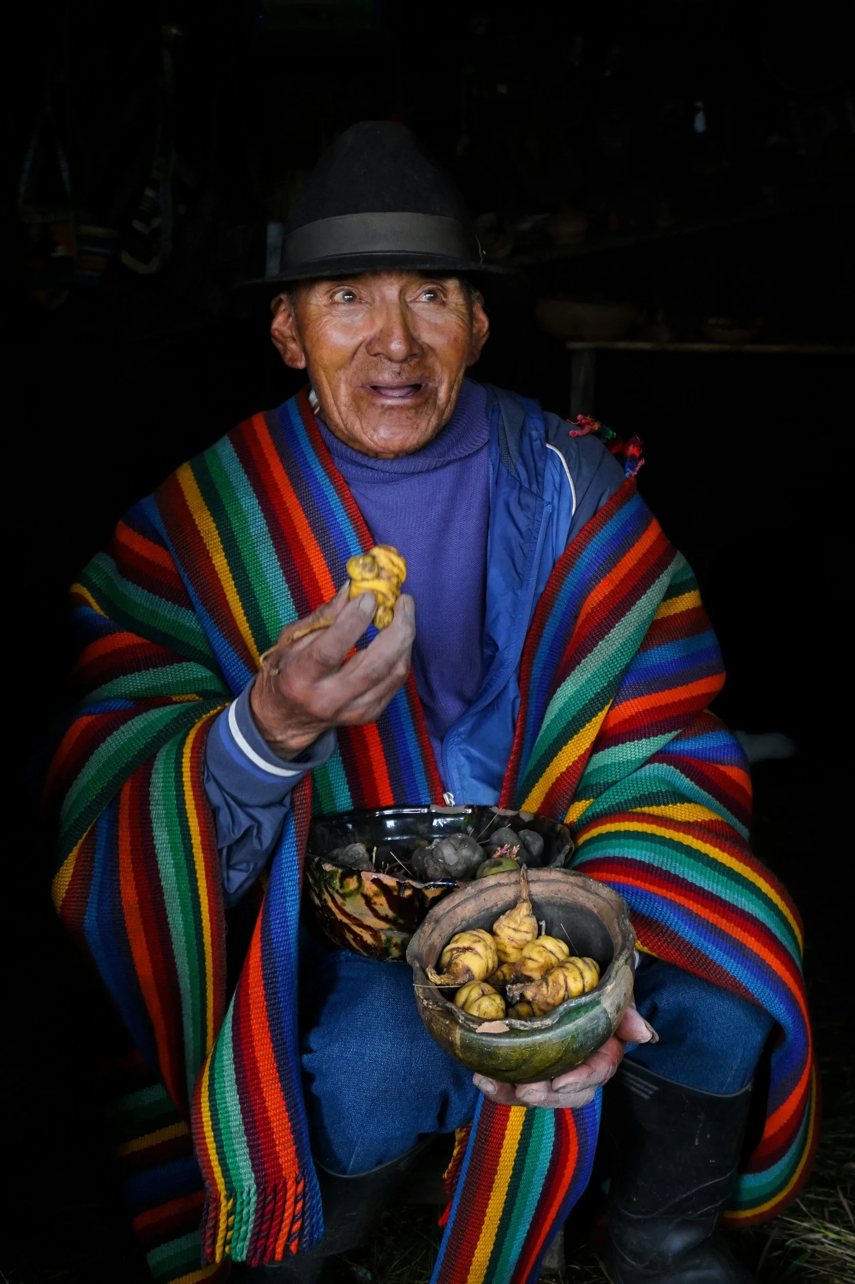 An elderly person wearing a colorful striped shawl and a black hat, holding a bowl of yellow and brown fruit or tubers, with a surprised or amused expression on their face.