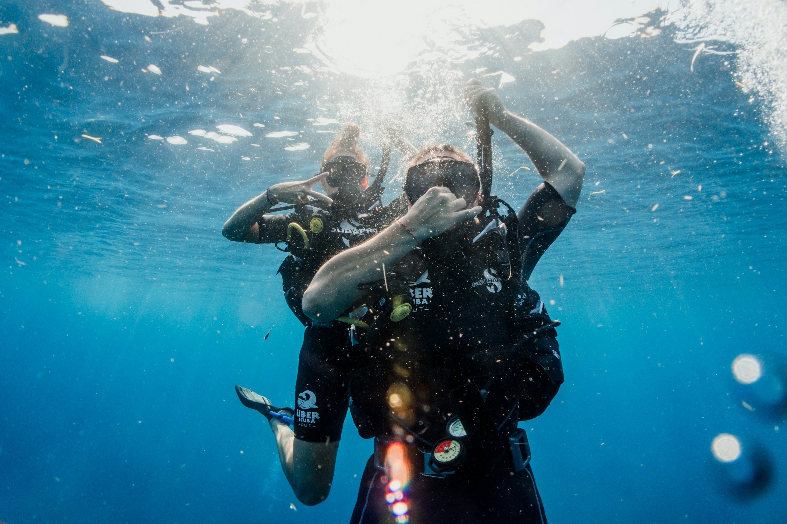 Two scuba divers underwater making peace signs with their hands, wearing masks and wetsuits, with sunlight filtering through the water above.