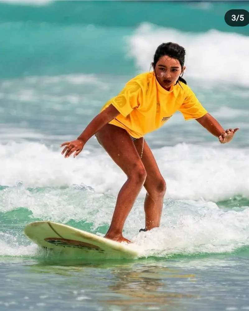 A girl in a yellow shirt stands on a surfboard riding a wave in the ocean.