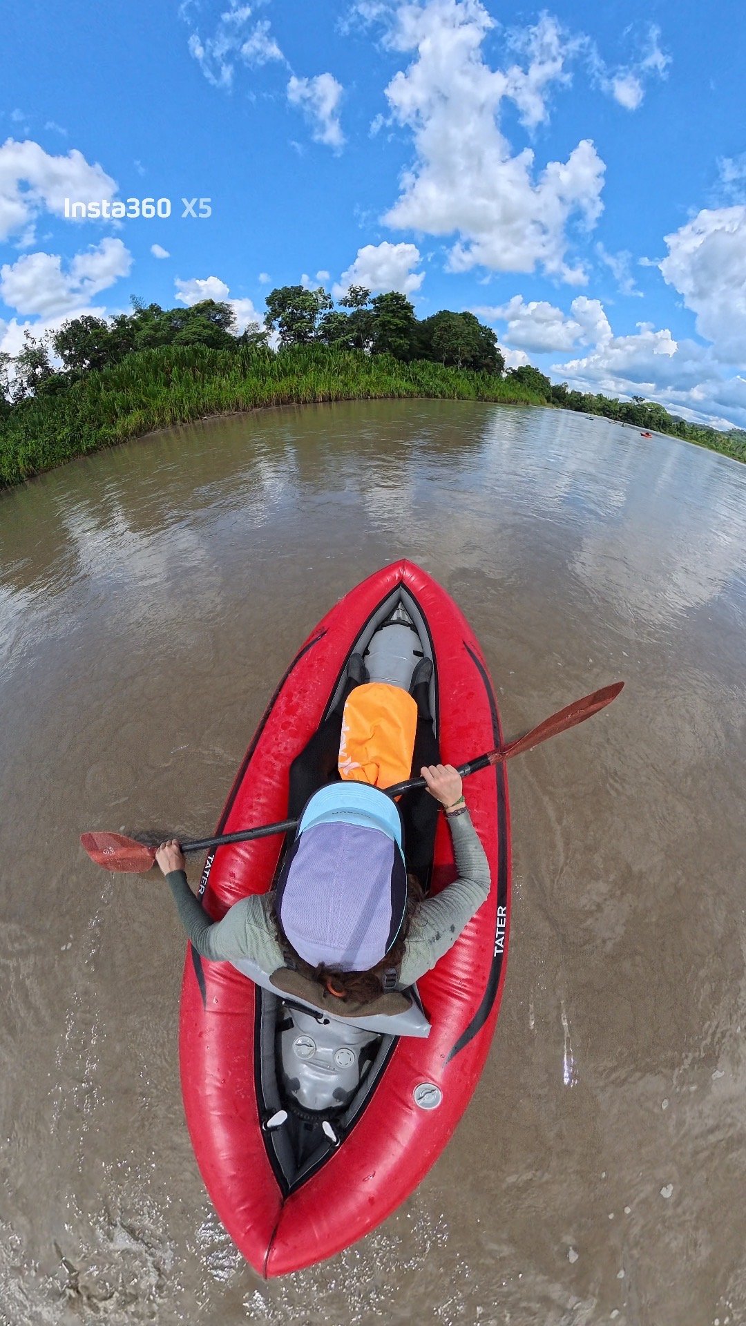 Top-down view of a person kayaking on a river under a cloudy sky with greenery along the riverbanks.