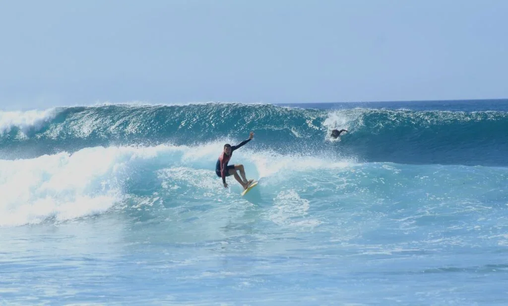 A person surfing a large wave with another surfer visible further back in the ocean.
