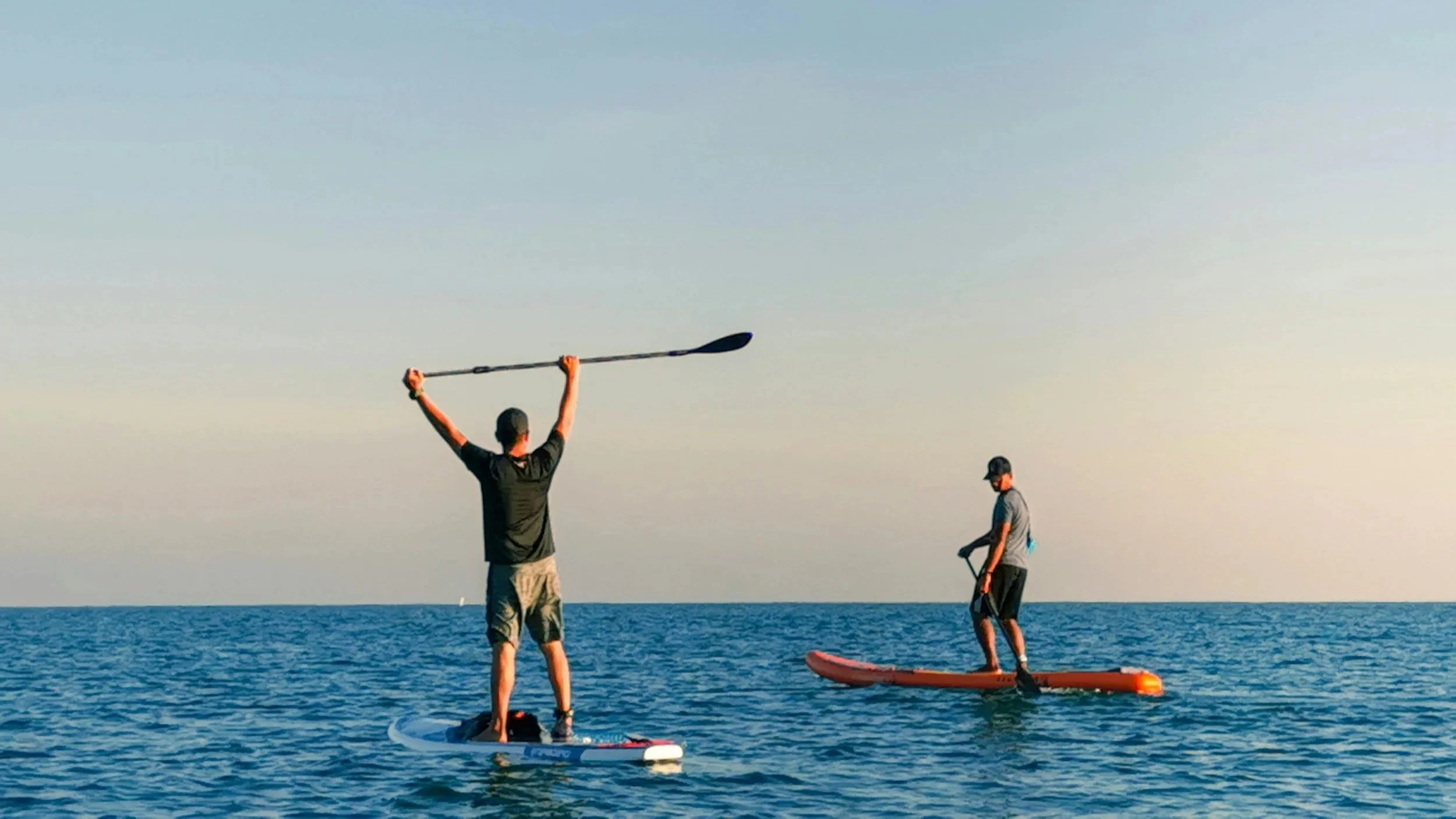 Two men paddleboarding on the ocean during sunset, with one man standing and holding a paddle raised above his head and the other man kneeling on his paddleboard.