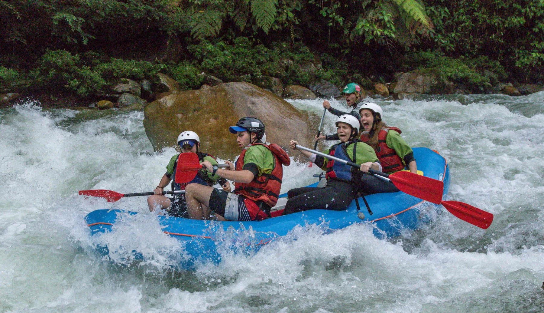 Group of five people wearing helmets and life jackets white water rafting on a river with rocks and green foliage in the background.