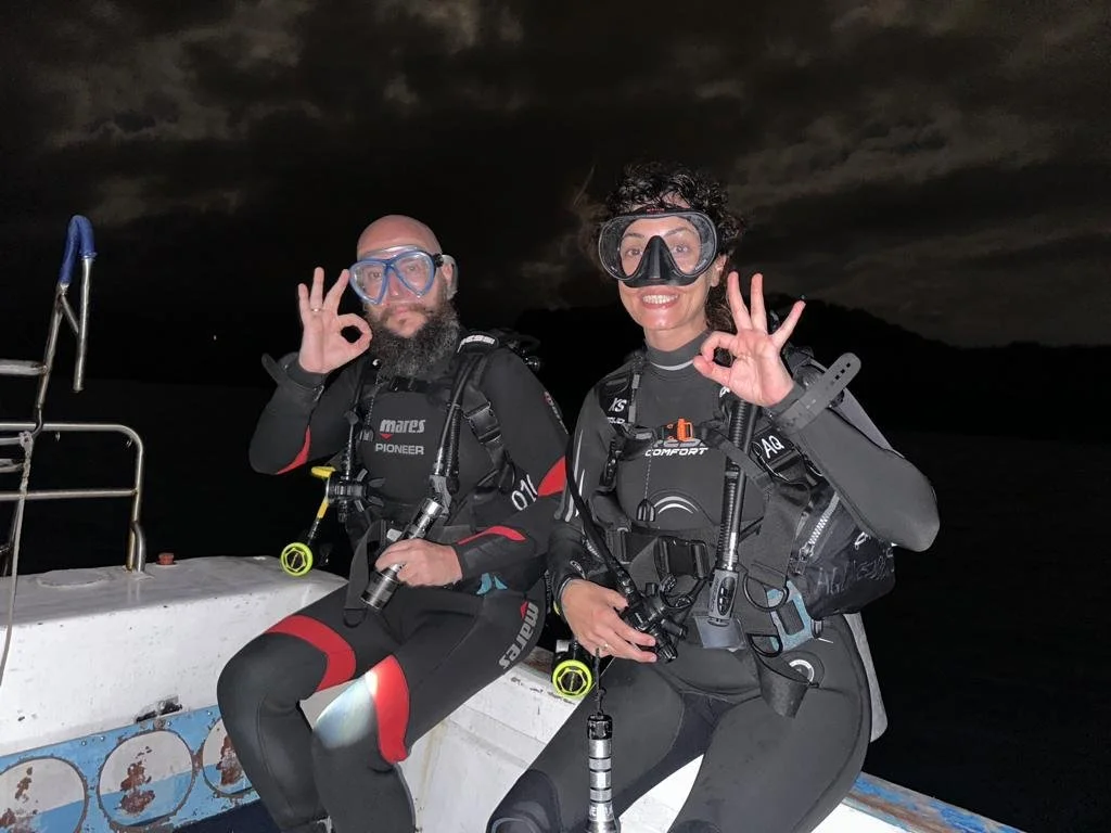 Two scuba divers in wetsuits on a boat at night, making OK signs with their hands, with dark water and cloudy sky in the background.
