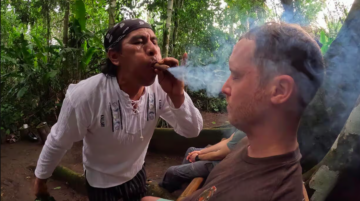 A man wearing a white shirt and black bandana smokes a cigar in a jungle setting with dense green foliage, while another man with short hair and a brown shirt sits with closed eyes nearby.