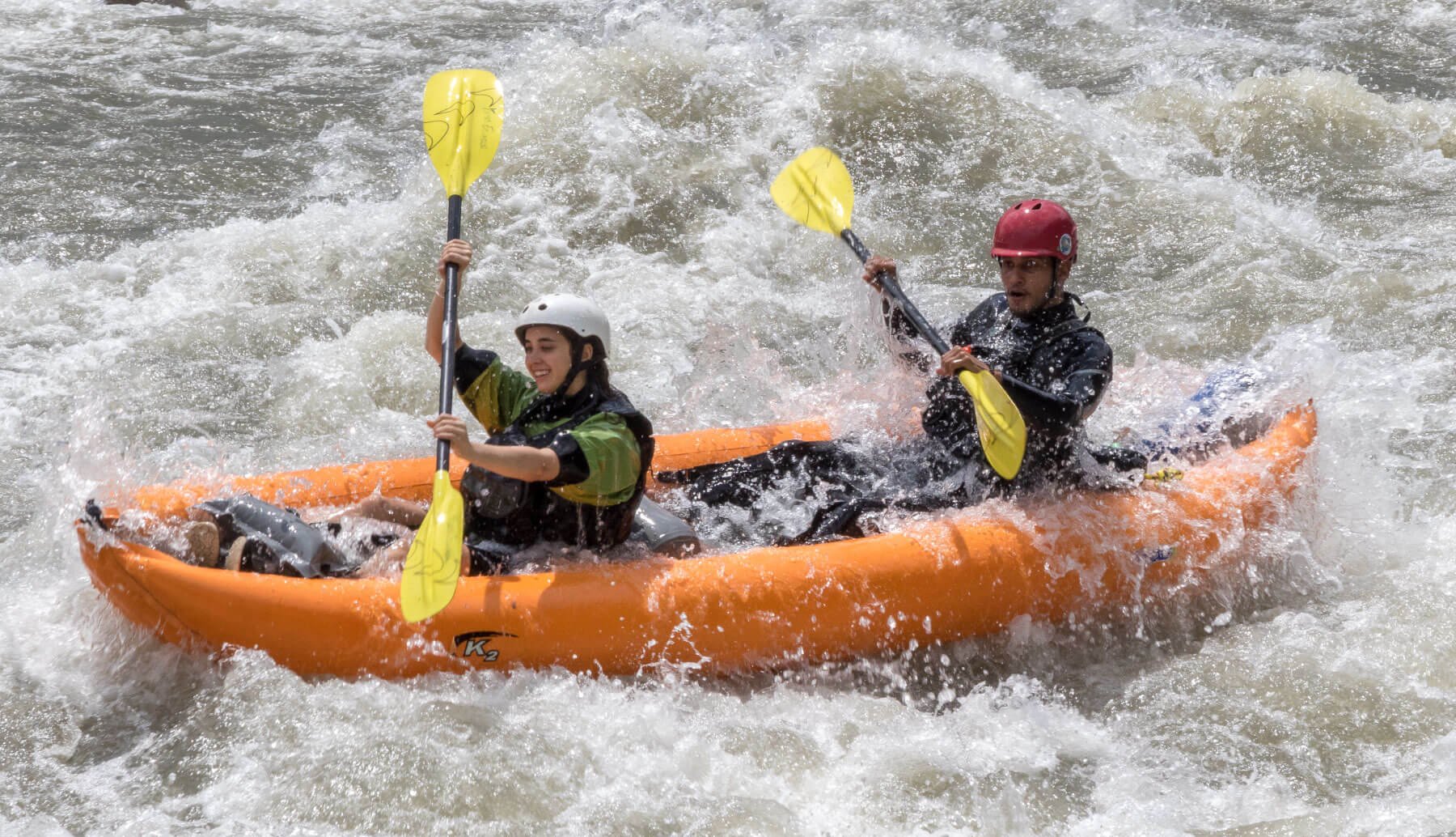 Two people white water rafting in an orange kayak, wearing life jackets and helmets, paddling through rough water with splashes around them.