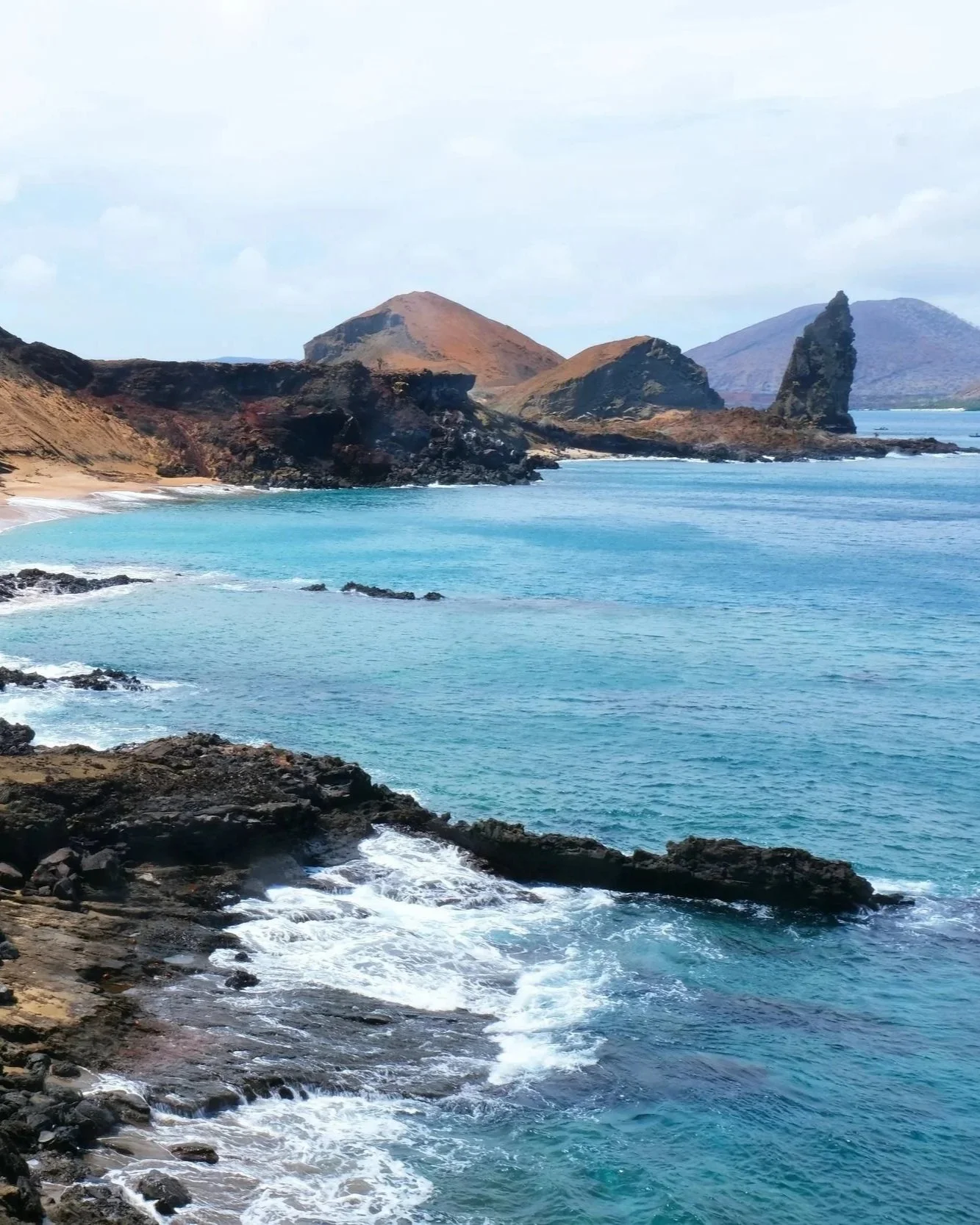 Scenic coastal landscape with rocky shoreline, turquoise ocean waters, and volcanic mountains in the background under a partly cloudy sky.