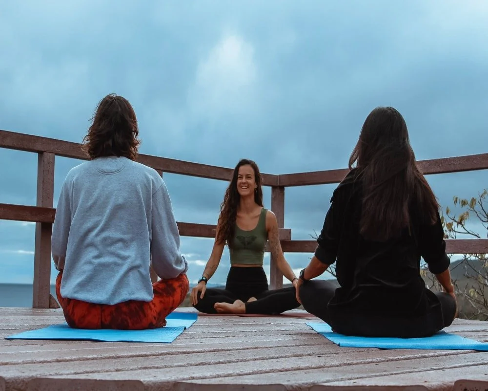 Three women practicing yoga on a wooden deck outdoors under a cloudy sky, with two women sitting cross-legged and the third woman sitting in a lotus position smiling.