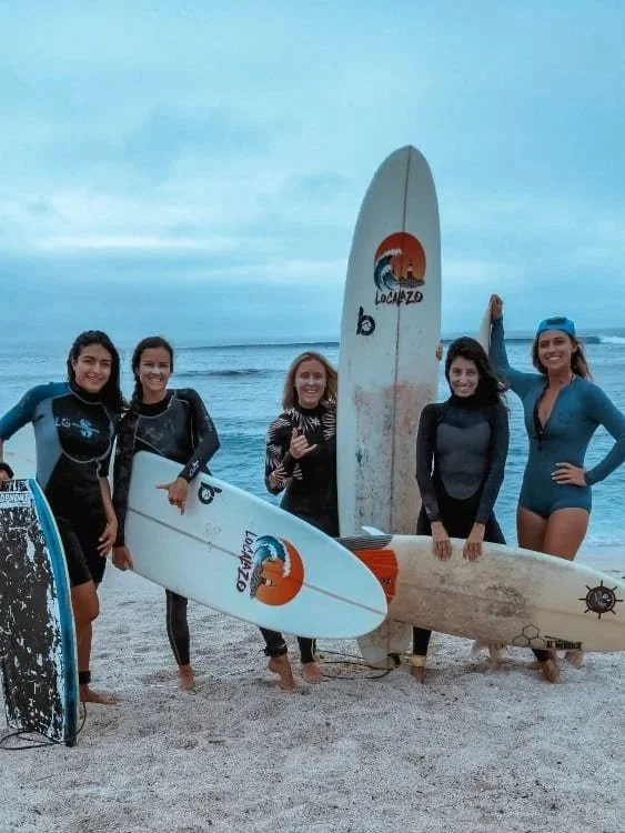 Five women standing on the beach with surfboards, smiling and posing for a photo.