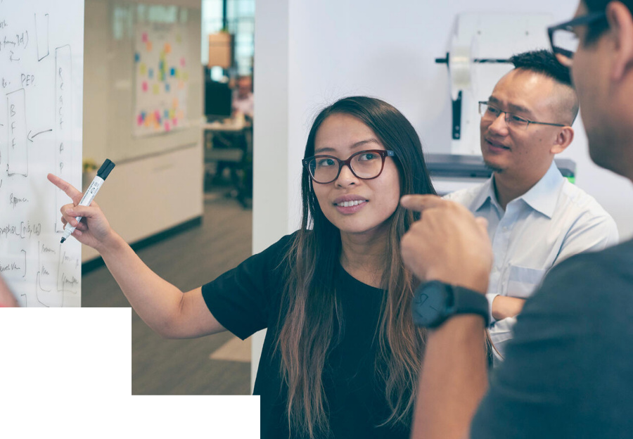 Three people having a discussion in an office, with one woman pointing at a whiteboard covered in notes and diagrams.