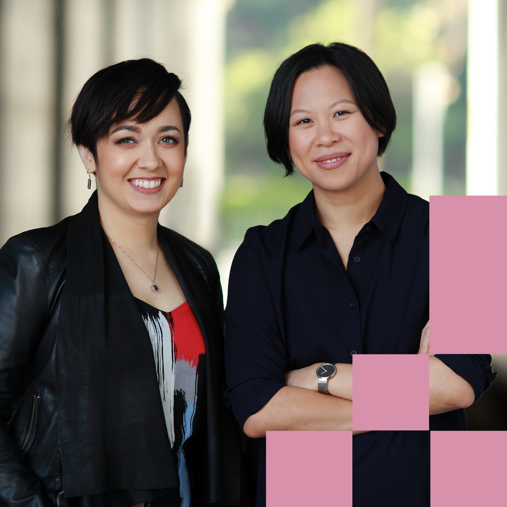 Two professional women smiling indoors, one in leather jacket, one in dark blue shirt, representing diverse tech team