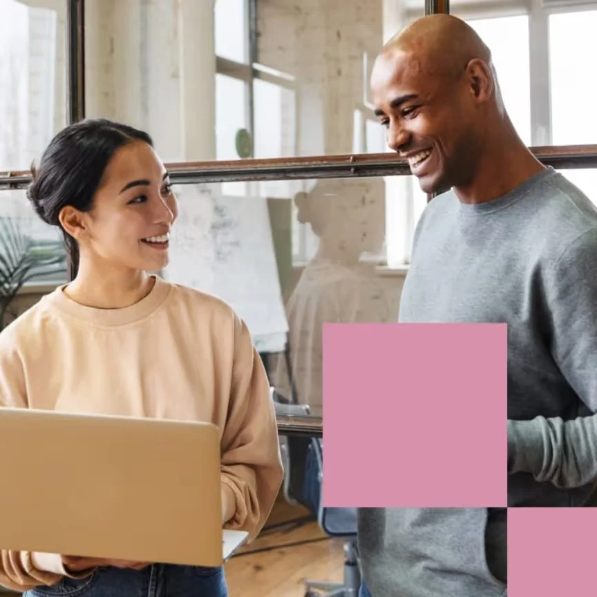 A young woman and a young man are having a conversation, smiling at each other in an indoor space with large windows.Young woman and man smiling while discussing work in inclusive tech office