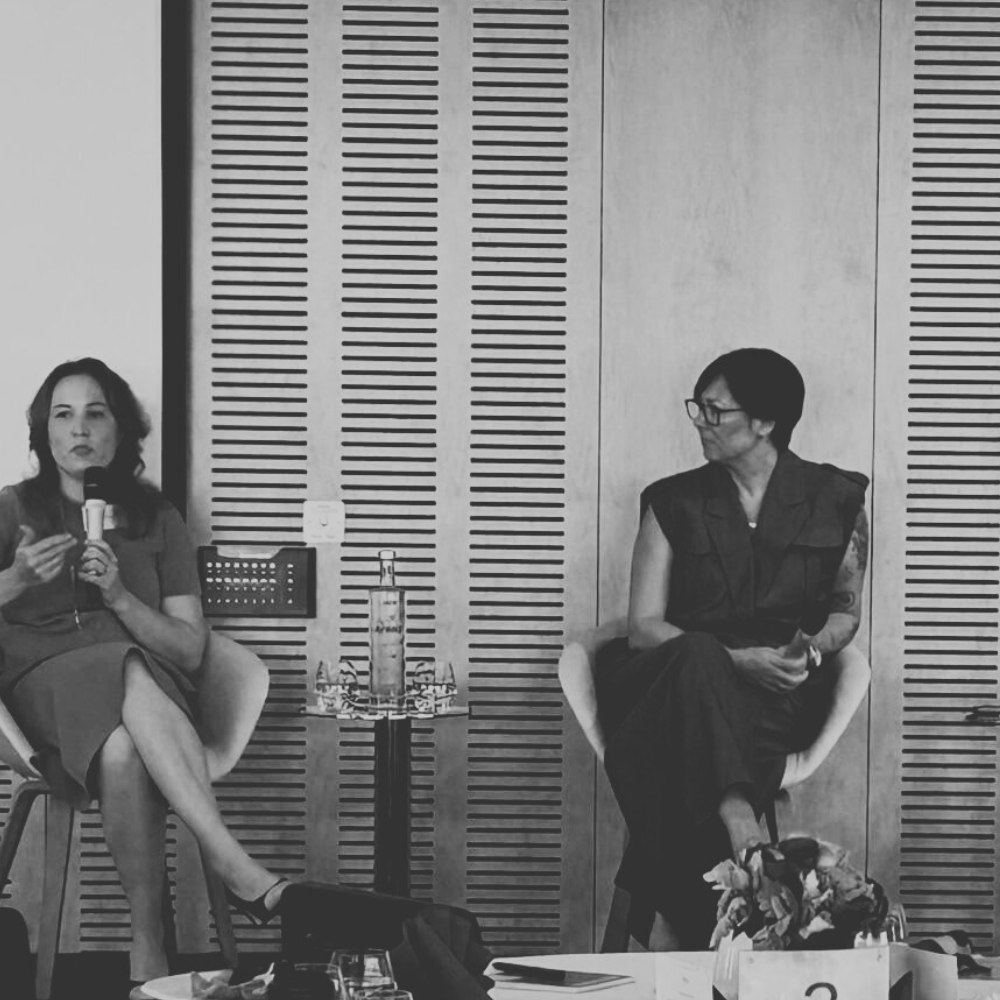 Two women speaking in panel discussion with table and flowers in room with wooden panels
