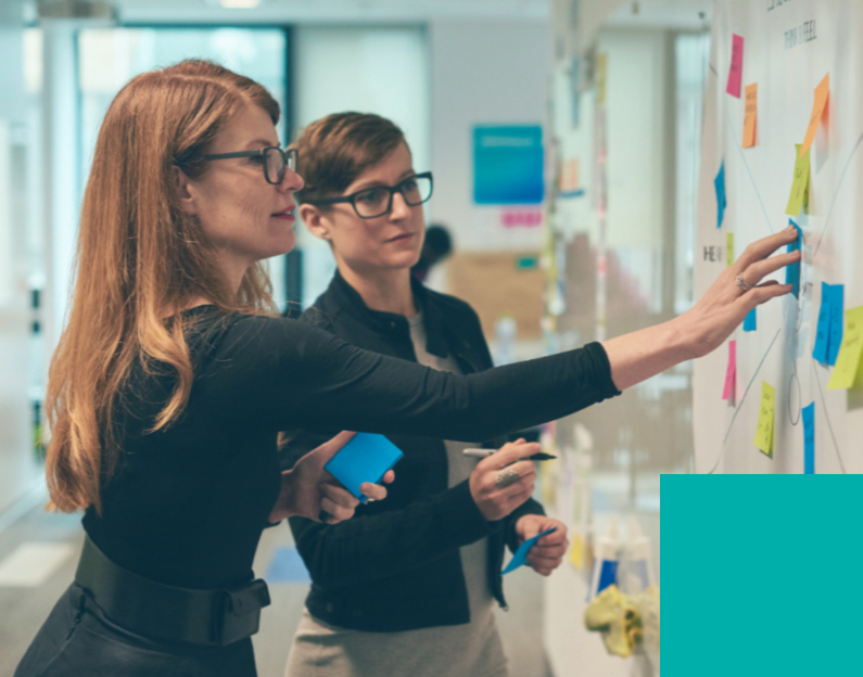 Two professional women with glasses collaborating and placing sticky notes on whiteboard in office
