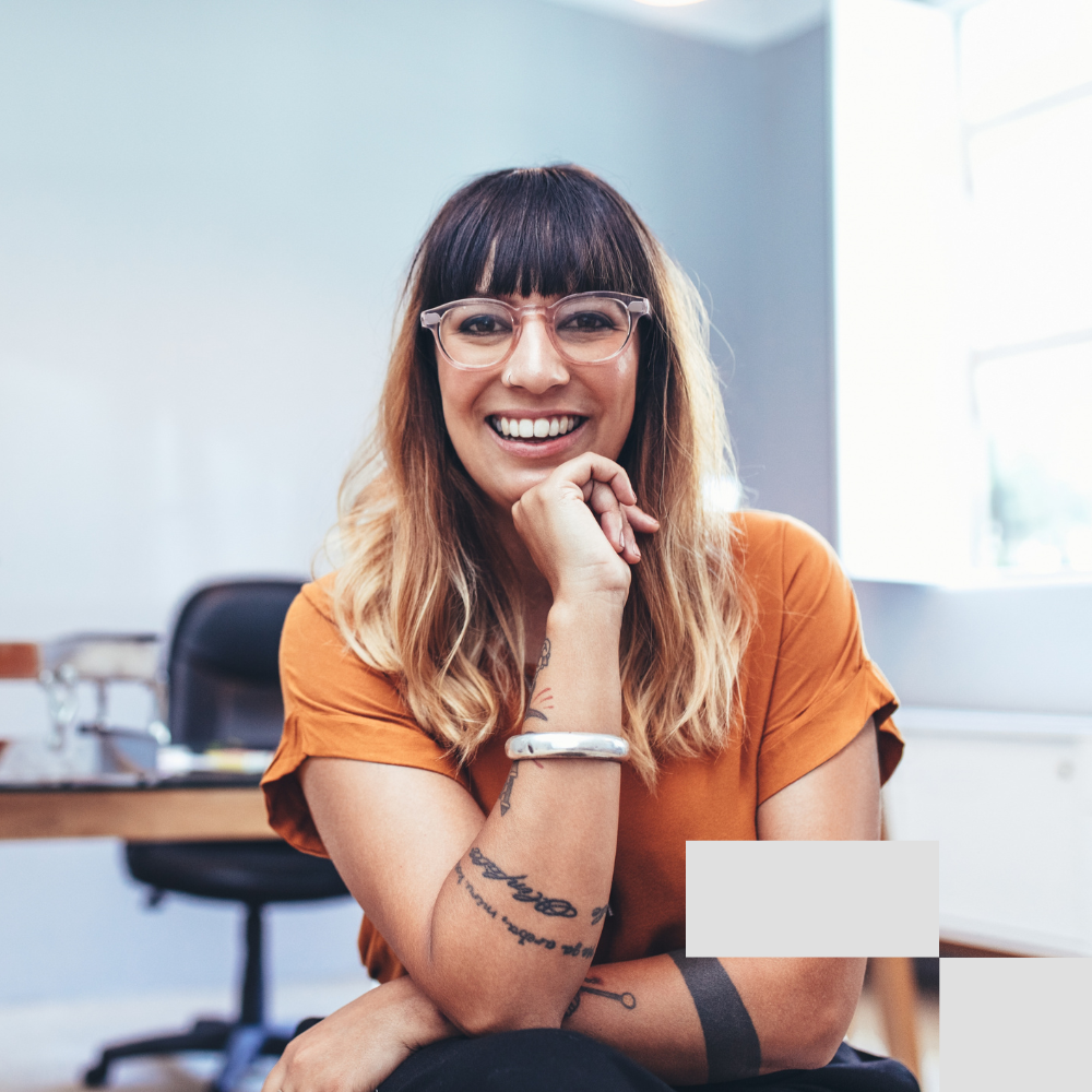 Smiling woman with glasses and tattoos sitting at desk in bright inclusive office