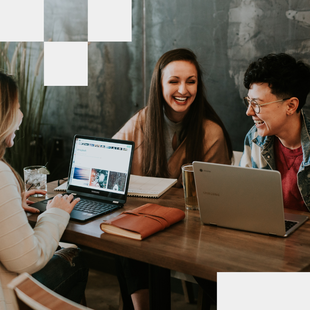 Three young women collaborating and laughing at wooden table using laptops in casual office setting