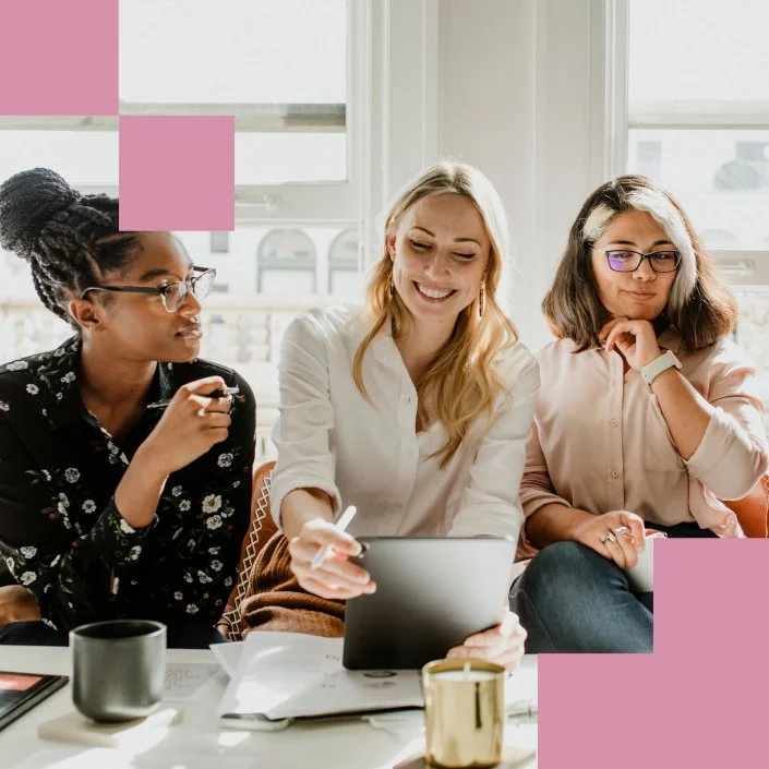 Three women collaborating at table using tablet in bright office with coffee mugs and papers