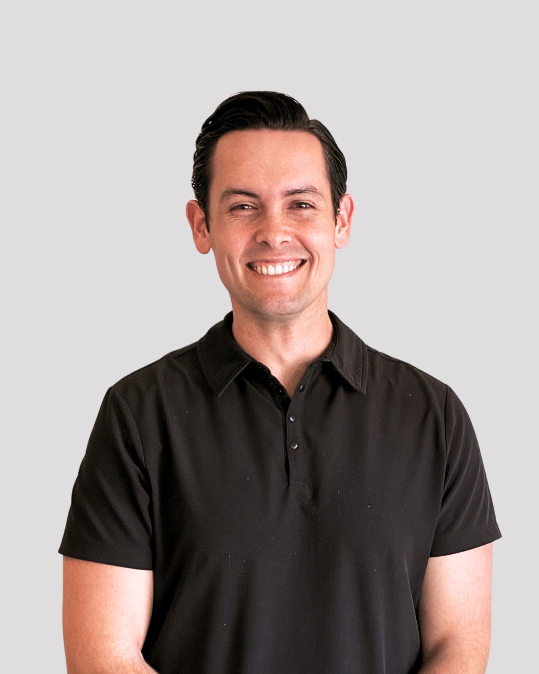 A man with dark hair, smiling, wearing a black polo shirt, standing against a plain light gray background.