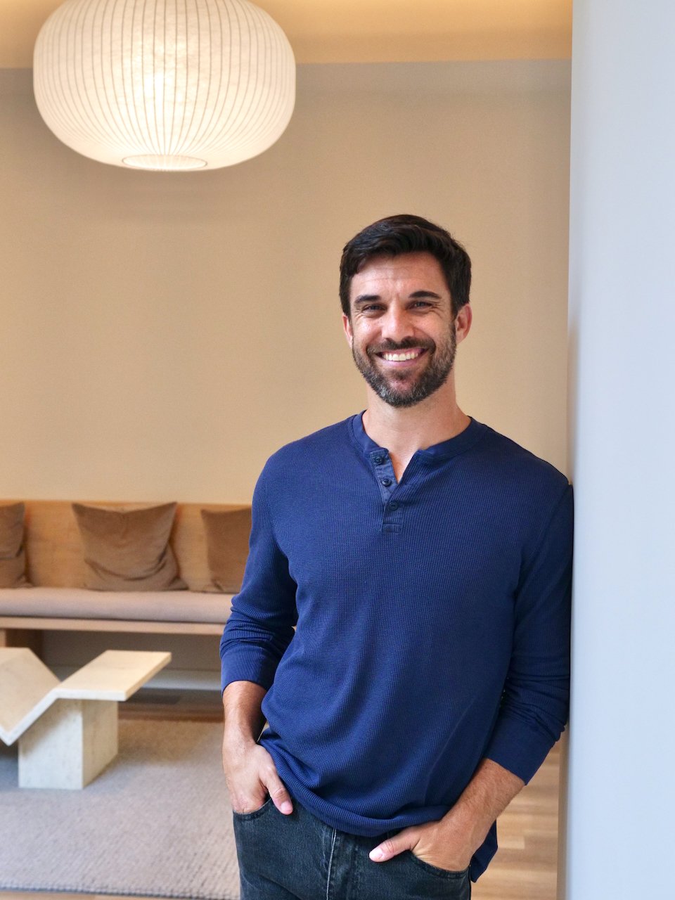 A man with dark hair and a beard smiling and leaning against a white wall in a living room with beige pillows on a couch and a large hanging lamp.