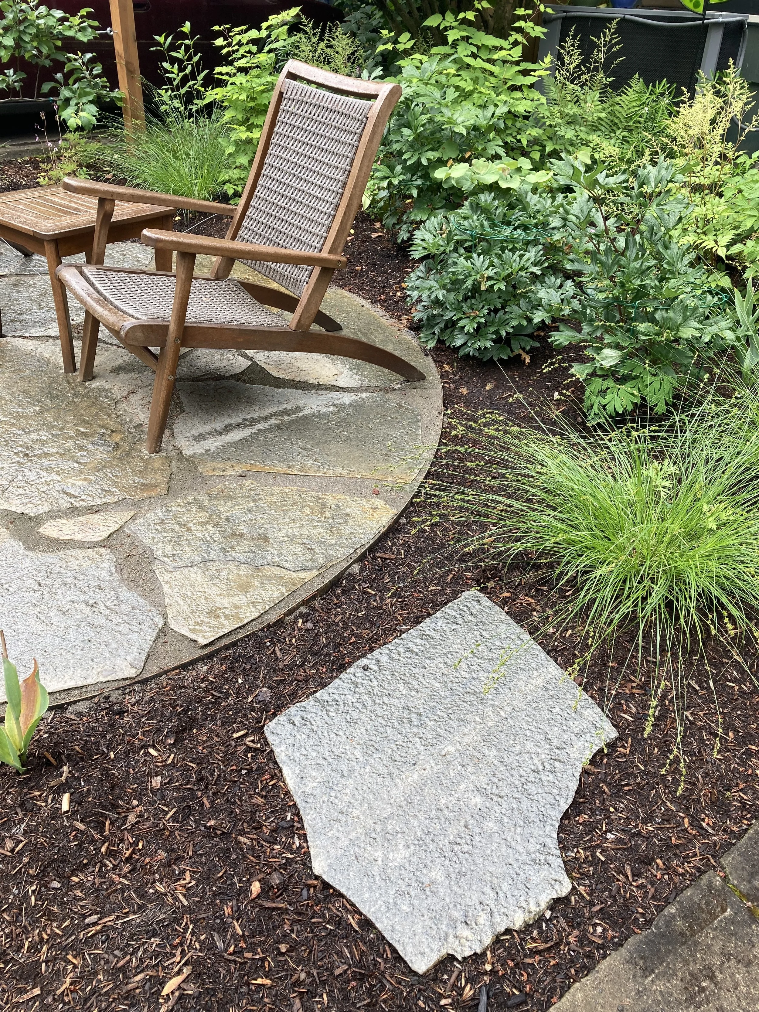 Wooden rocking chair and small table on a stone patio surrounded by green plants and mulch.