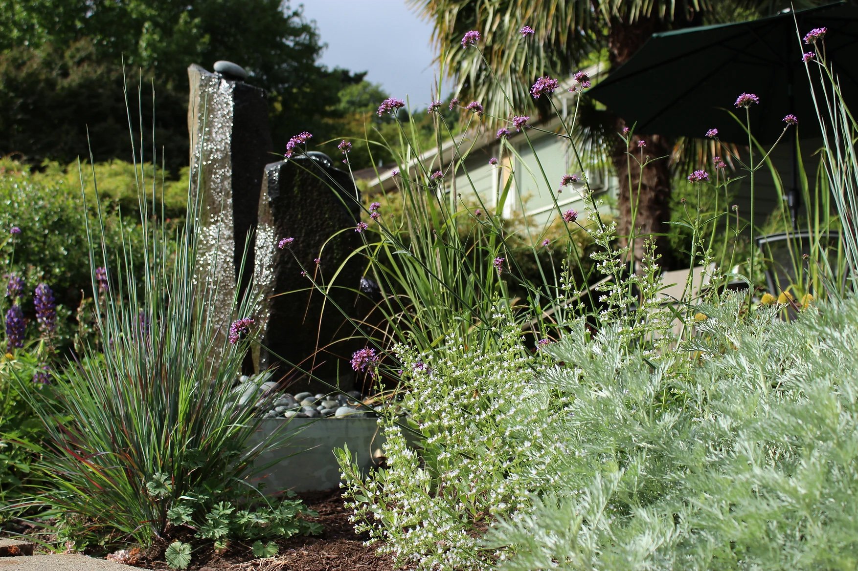A garden with various plants and flowers, a water fountain, and a shaded seating area with an umbrella.