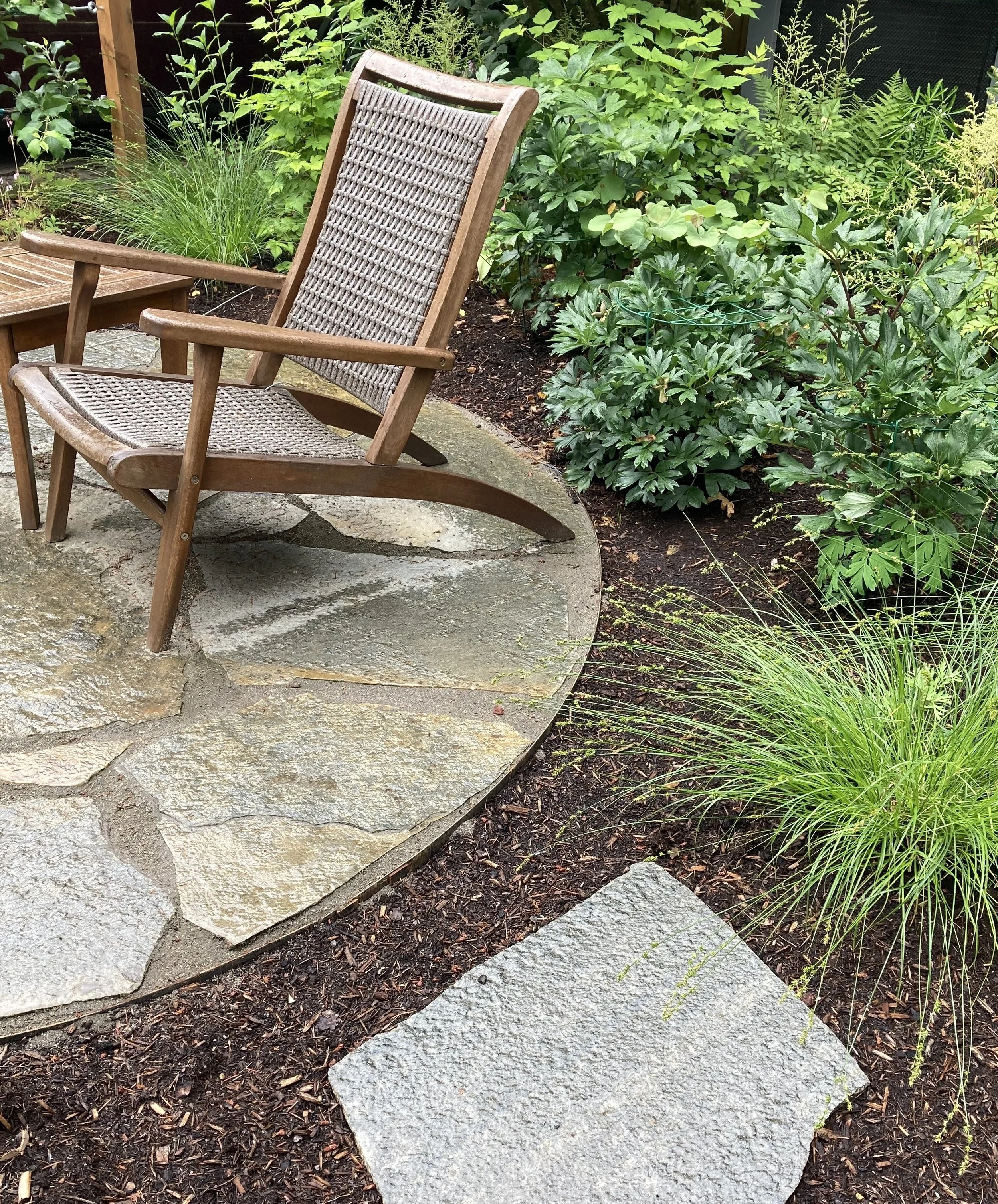 A wooden outdoor rocking chair on a stone patio surrounded by lush green plants and mulch.