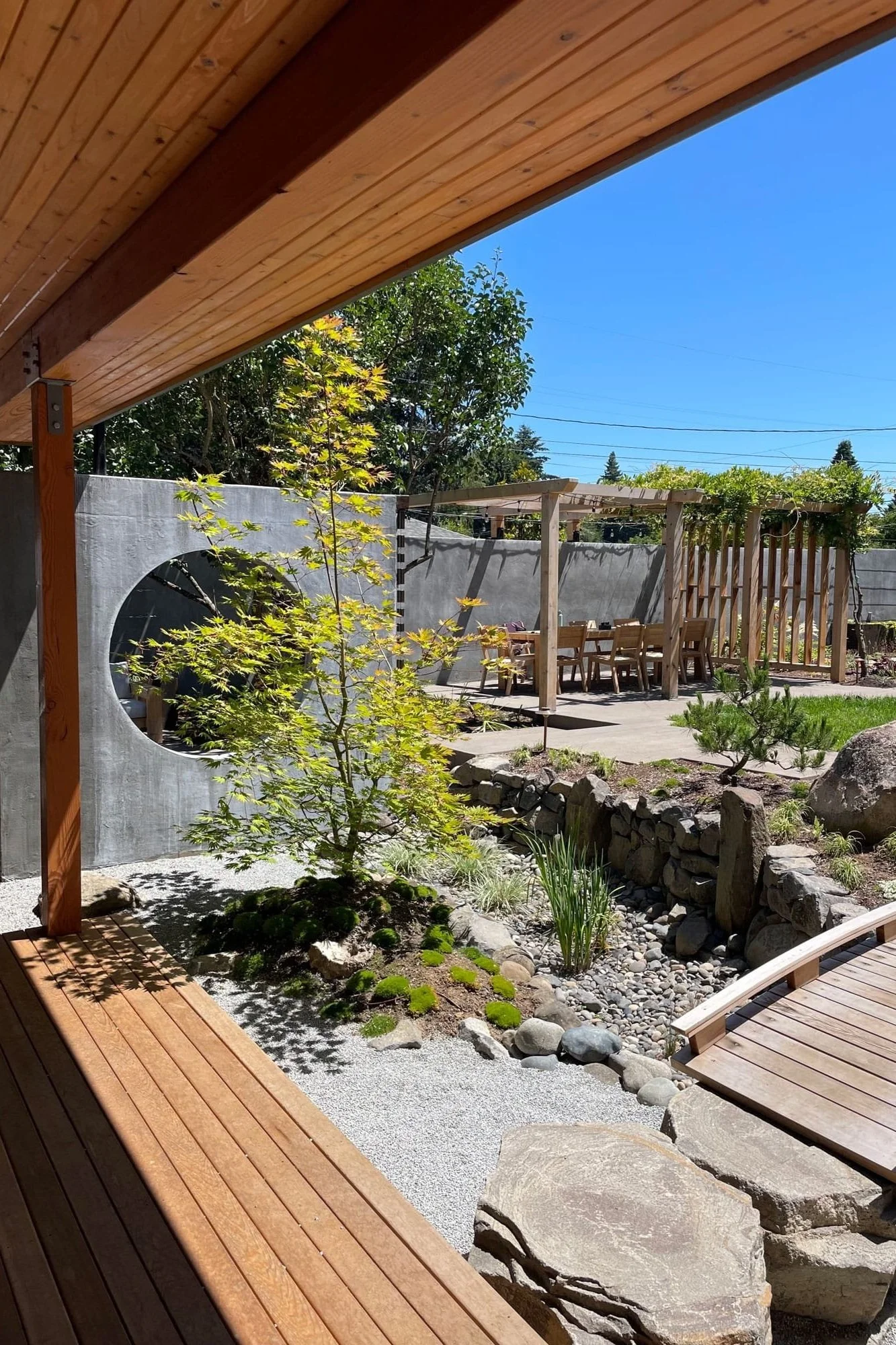 A backyard garden with a Japanese-style rock garden, small trees, and a wooden dining table with chairs under a pergola, with a wooden deck and shade structure in the foreground and a concrete wall with a circular cutout in the background.