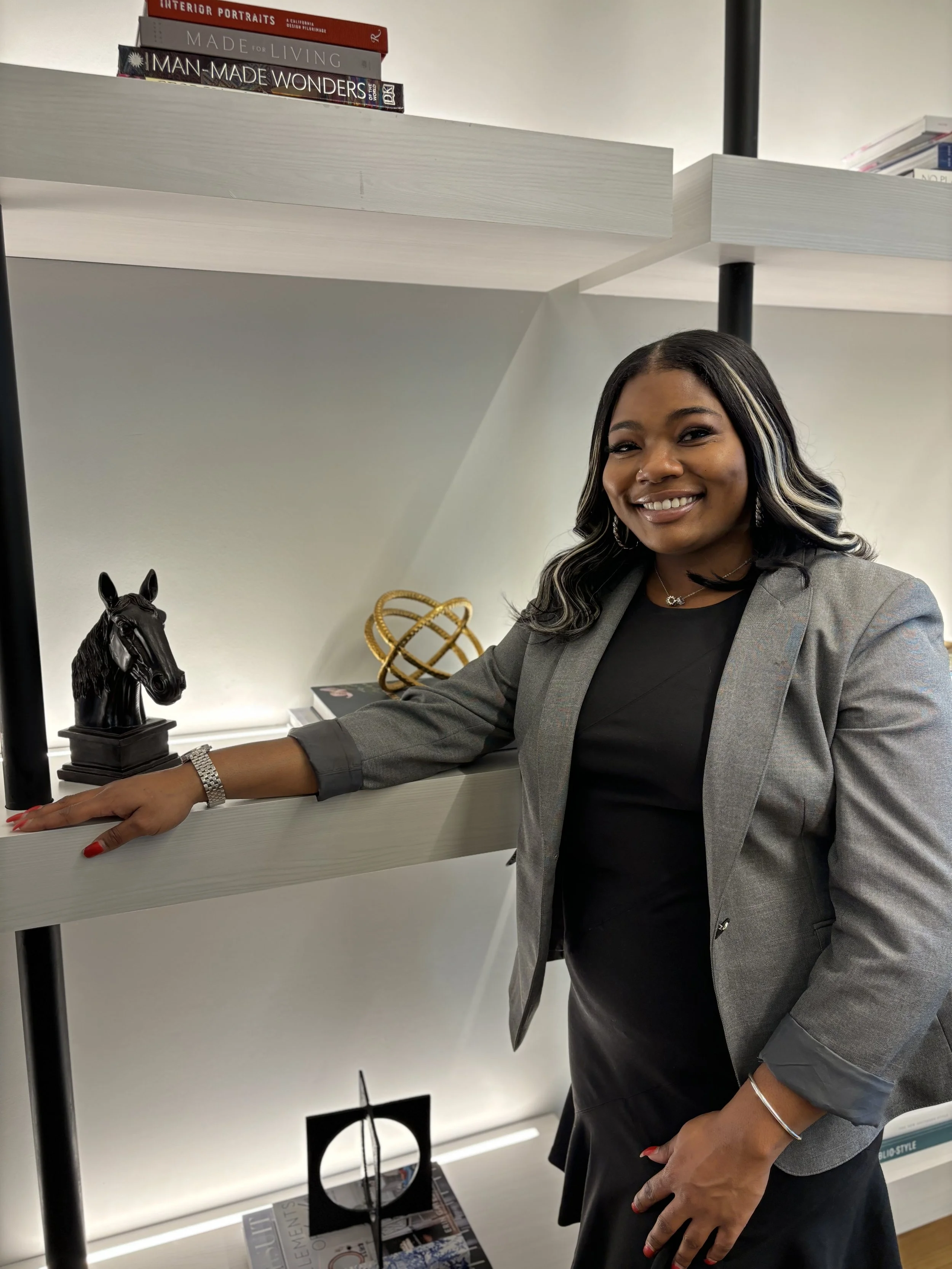 A woman with black hair and blonde highlights wearing a gray blazer and black dress, smiling, placing her hand on a bookshelf with decorative items including a black horse head sculpture and a gold geometric sculpture.