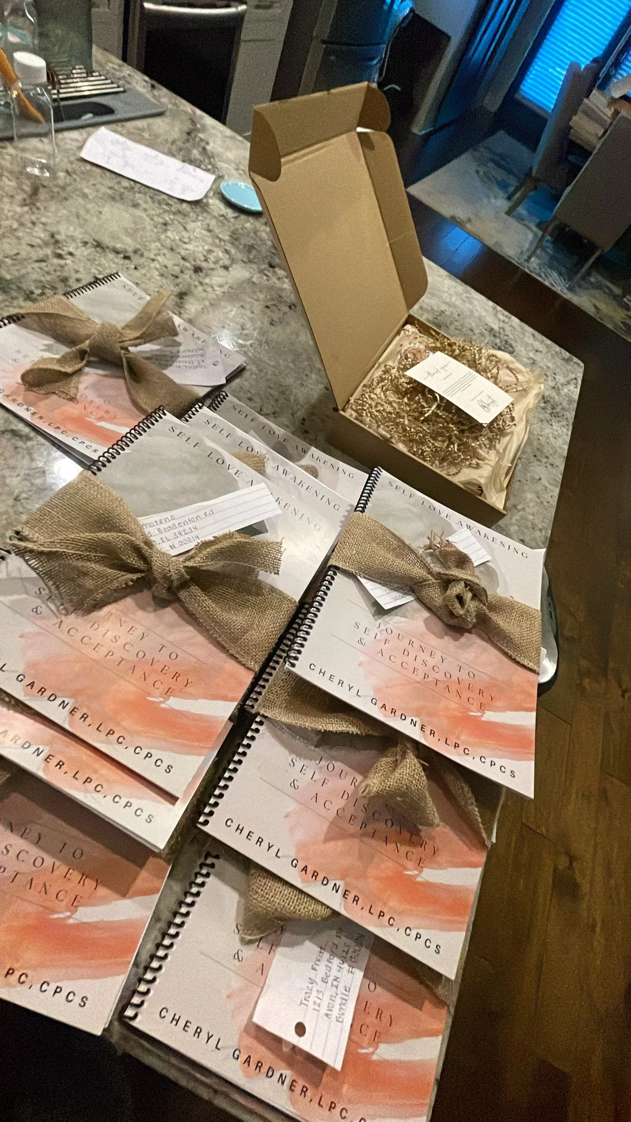 Multiple copies of a book titled "Self Love Awakening" by Cheryl Gardner, each wrapped with a burlap bow, arranged on a kitchen counter, with an open box and a notepad nearby.