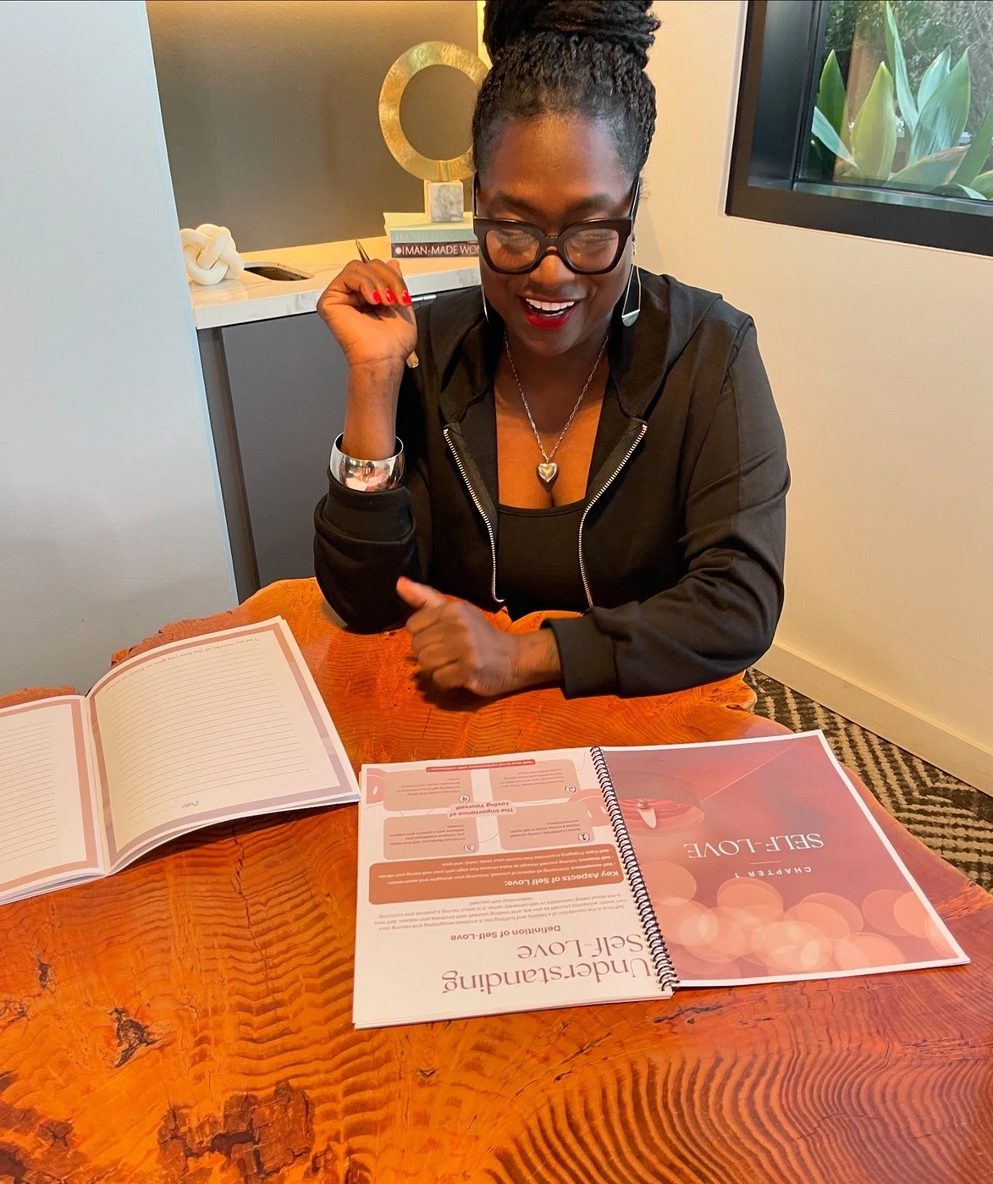 A woman with glasses, wearing a black jacket, sitting at a wooden table with a cheerful expression. She has a necklace with a heart pendant, and her hair is styled in braids. On the table are a notebook, a binder titled 'Understanding Self-Love,' and an open lined notebook. In the background, there is a window with green plants visible outside and a decorative round mirror on a gray and white cabinet.