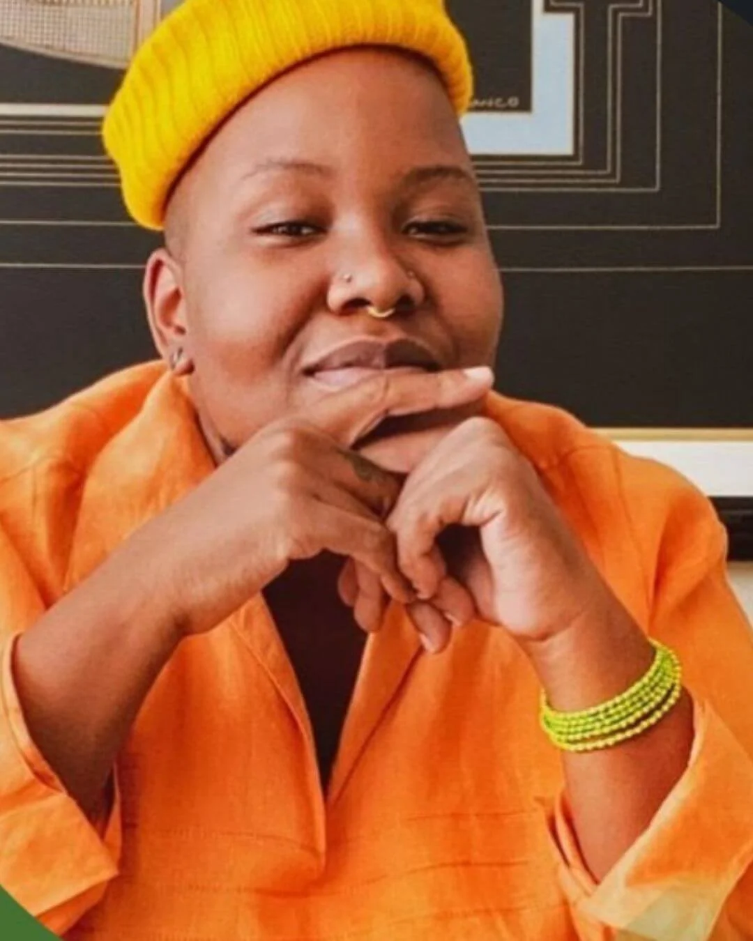 A person with a dark skin tone wearing a yellow knit cap, orange shirt, and yellow beaded bracelet, resting their chin on their hands in a relaxed pose.