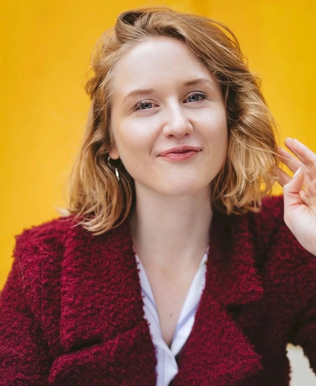 A woman with wavy blonde hair and light skin smiling gently, wearing a dark red textured coat and a white shirt, standing against a yellow background.