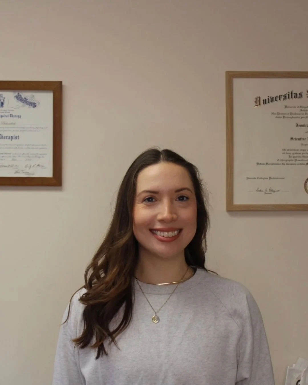 A woman with long brown hair and a light gray shirt smiling in front of a beige wall with framed diplomas.